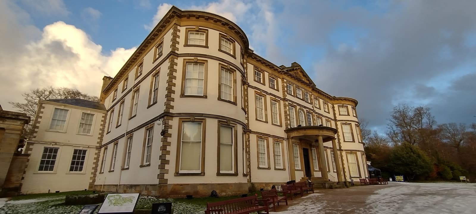 A grand, three-story historic building with a rounded corner and large windows sits against a backdrop of a partly cloudy sky. The surrounding area has light snow, and there are benches and an information board on the path in front.