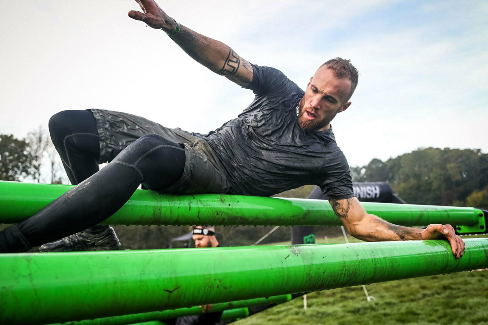 A man in athletic gear and knee-high socks maneuvers over bright green horizontal bars in an outdoor obstacle course. He appears to be in mid-motion, balancing himself with one hand while swinging his leg over. The background features trees and a blurred participant.