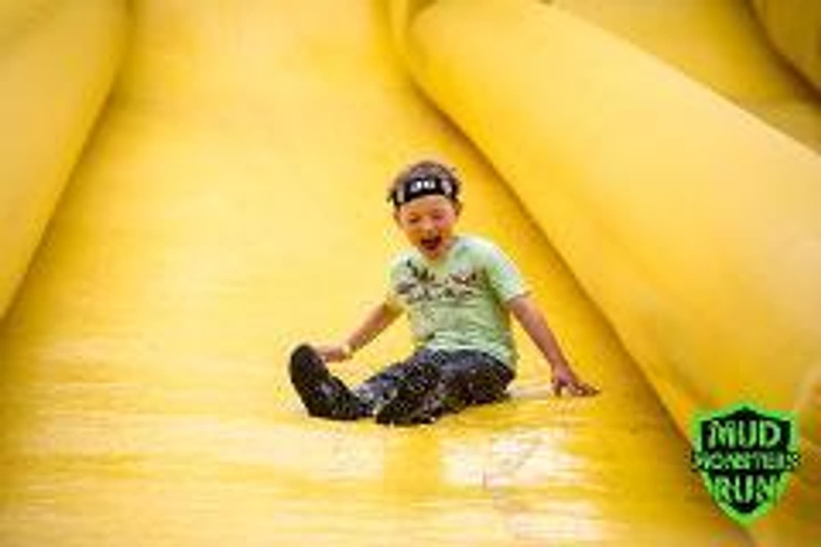 Child wearing a headband and green shirt slides down a large yellow inflatable slide with a joyful expression. The slide is part of a Mud Monsters Run event, indicated by the logo in the corner.