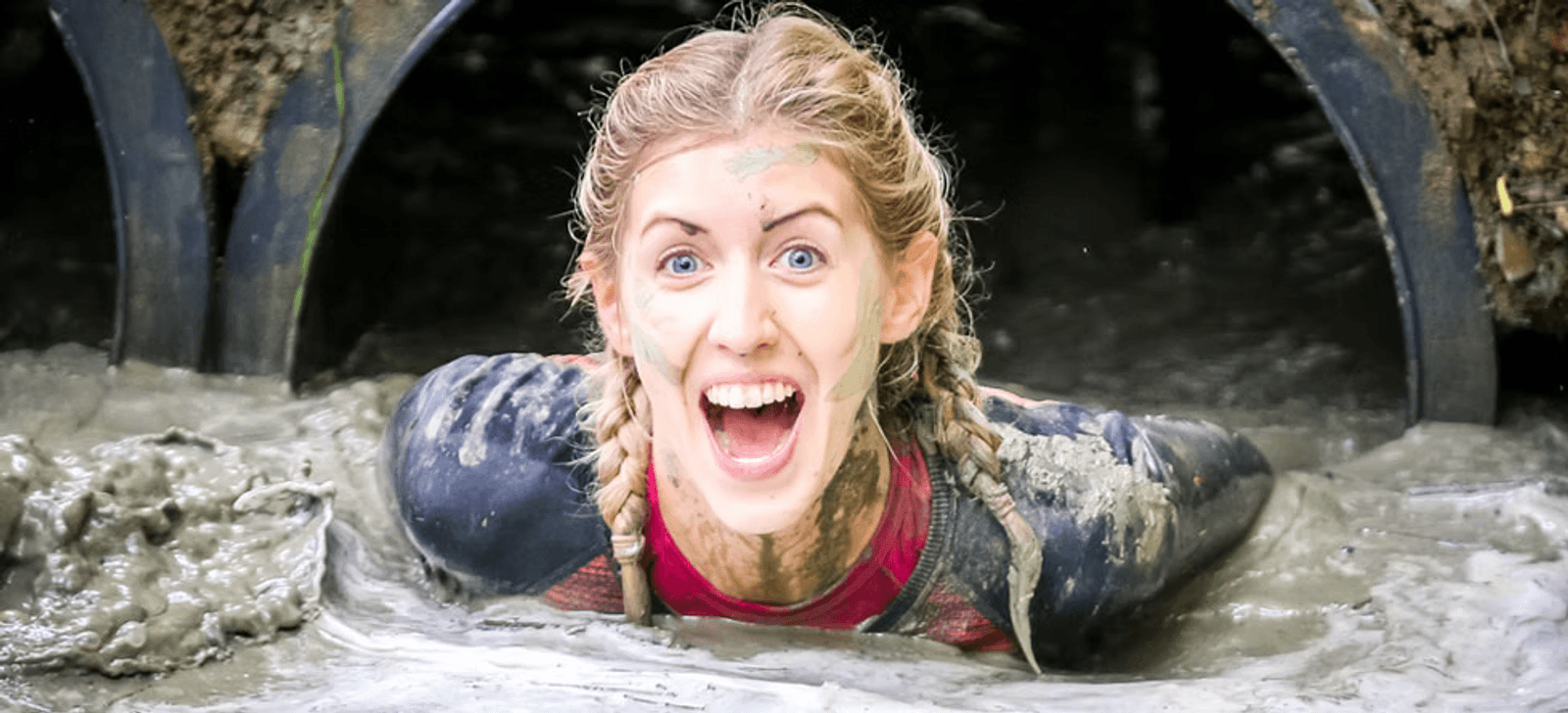 A person with braided hair, covered in mud, emerges excitedly from a tunnel during an obstacle course. They are wearing a long-sleeve athletic top and appear joyful, with mud splattered on their face and clothes.
