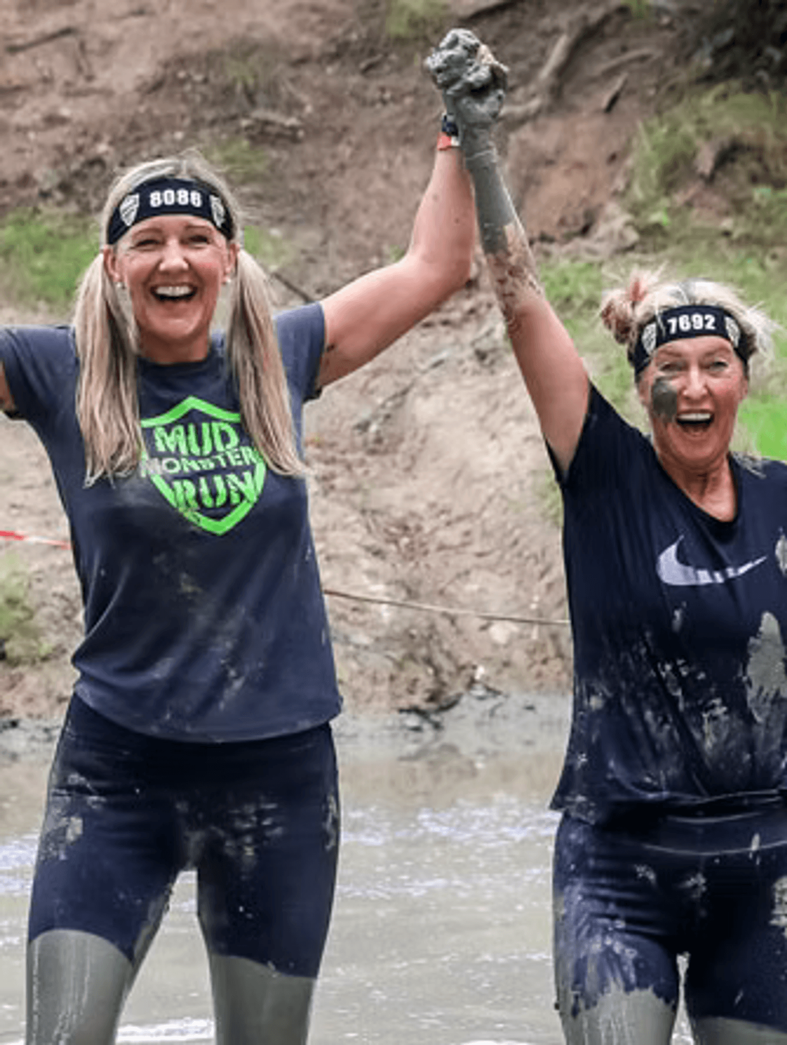 Two women covered in mud celebrate with arms raised and smiles after a mud run. They wear black shirts and headbands with numbers, standing in front of a muddy obstacle course.