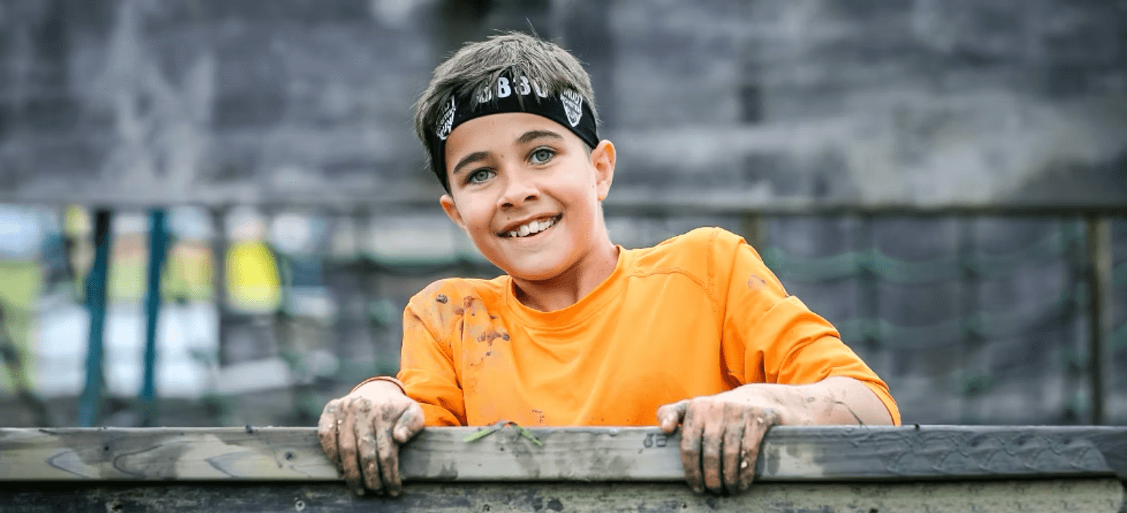 A smiling child with short brown hair and a black headband, wearing an orange shirt, leans over a wooden obstacle. His hands and shirt are muddy. The background is blurred, showing more obstacles and greenery.