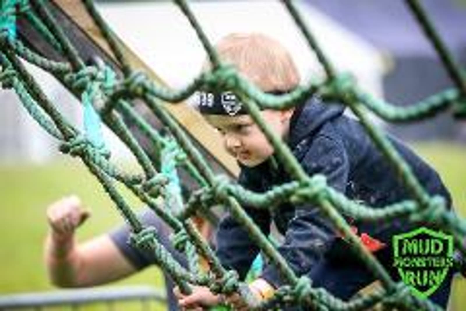 Young child wearing a headband climbs a rope net during an outdoor obstacle course event. The child is focused and wears a dark jacket. Part of an adult's arm is visible, and there's a "Mud Monsters Run" logo in the corner.