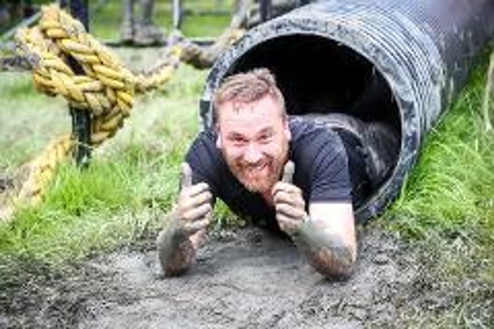A man covered in mud crawls out of a muddy pipe on an obstacle course, giving two thumbs up. The grimace on his face looks like he's exerting a lot of effort, and yellow ropes can be seen on the left side.