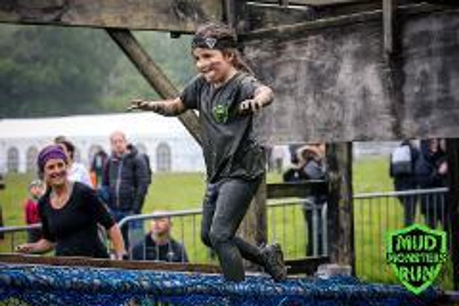 A person covered in mud balances on a narrow wooden beam during an outdoor obstacle course. Onlookers stand behind a fence, and a tent is visible in the background. The image has a "Mud Monsters Run" logo in the corner.