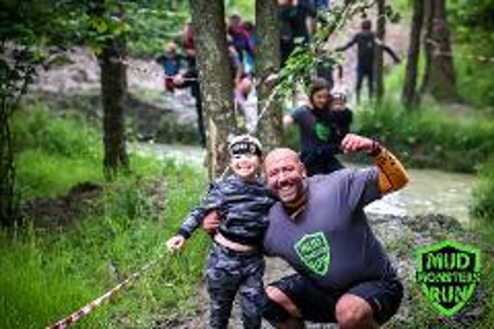 A man and a child, both muddy and smiling, pose proudly in front of a muddy obstacle run course. The man flexes his arms while the child holds a thumbs up. People in the background navigate the muddy trail and stream. Trees surround the event.
