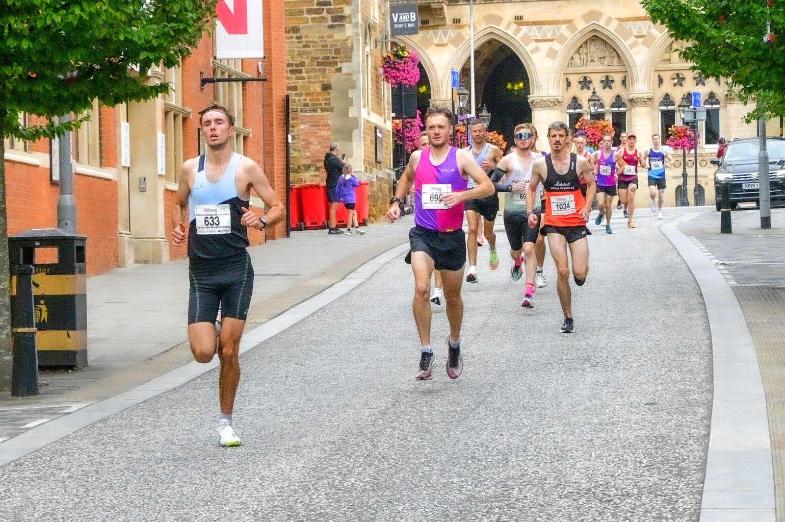 Runners participating in a city marathon ascend a slight incline. The leading runner wears a white tank top and black shorts while others follow close behind. Buildings with flower decorations line the path, and trees provide shade along the street.