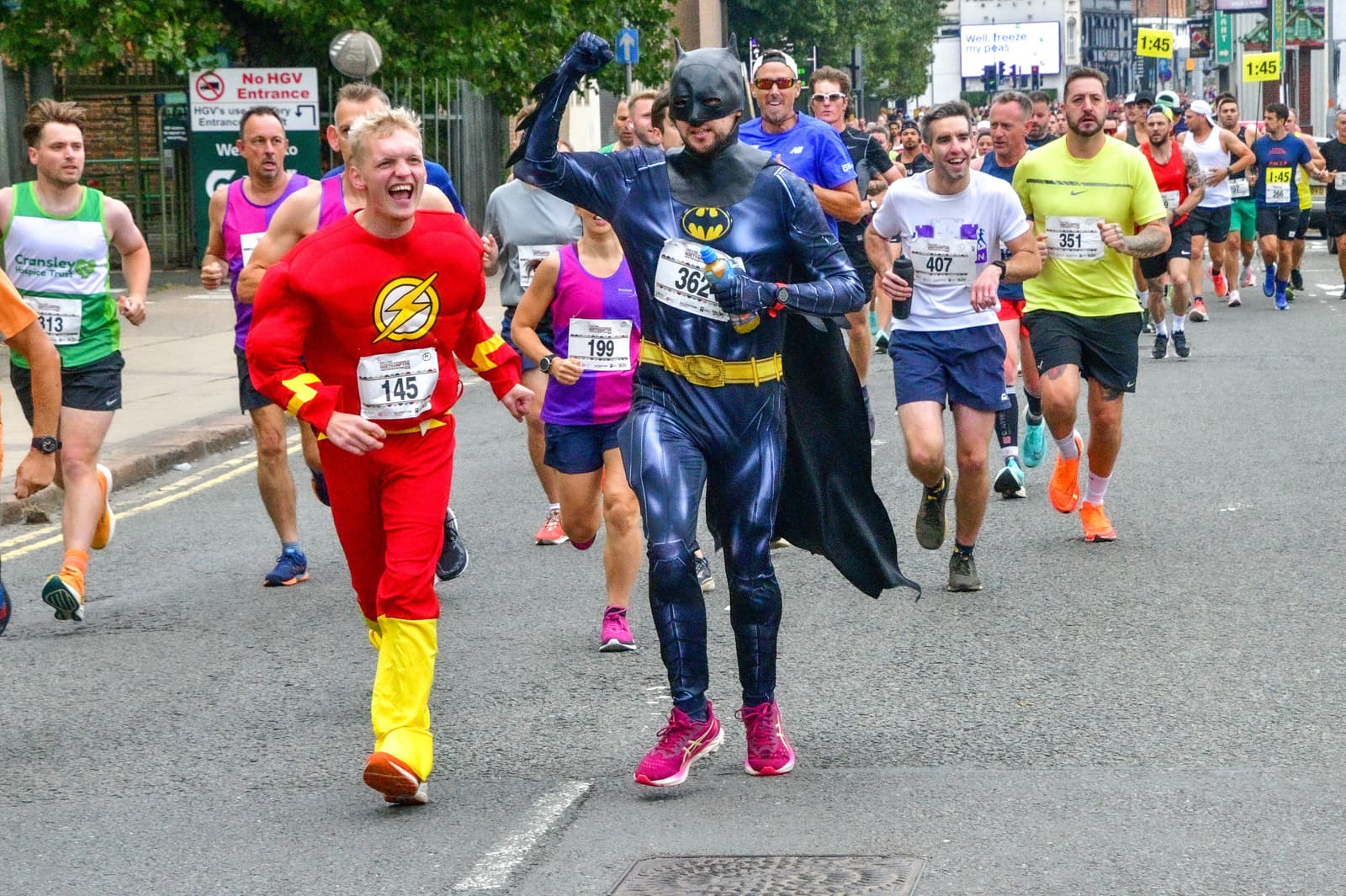 A group of runners participating in a race, with two of them dressed in superhero costumes: one as The Flash (red suit with a lightning bolt) and one as Batman (dark suit and cape). They're surrounded by other runners in athletic gear on a city street.
