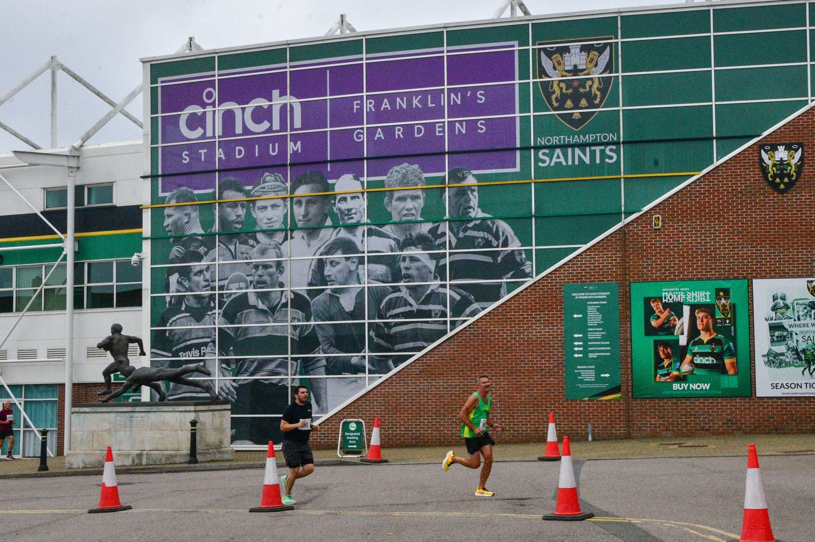 Exterior of Franklin's Gardens stadium, prominently featuring a large mural with rugby players, and the signage for Cinch Stadium and Northampton Saints. In front of the stadium, two individuals are running past traffic cones.