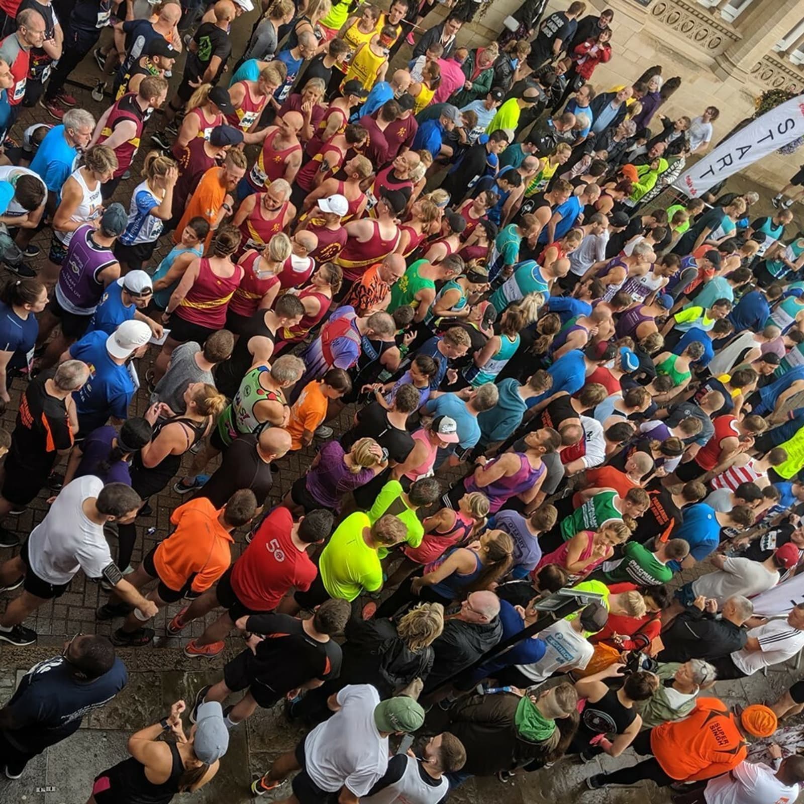 A large crowd of runners, dressed in colorful athletic gear, gather at the start line of a race. The scene is lively and packed, with participants standing closely together, awaiting the beginning of the event. Brightly colored shirts and hats are prominent in the crowd.
