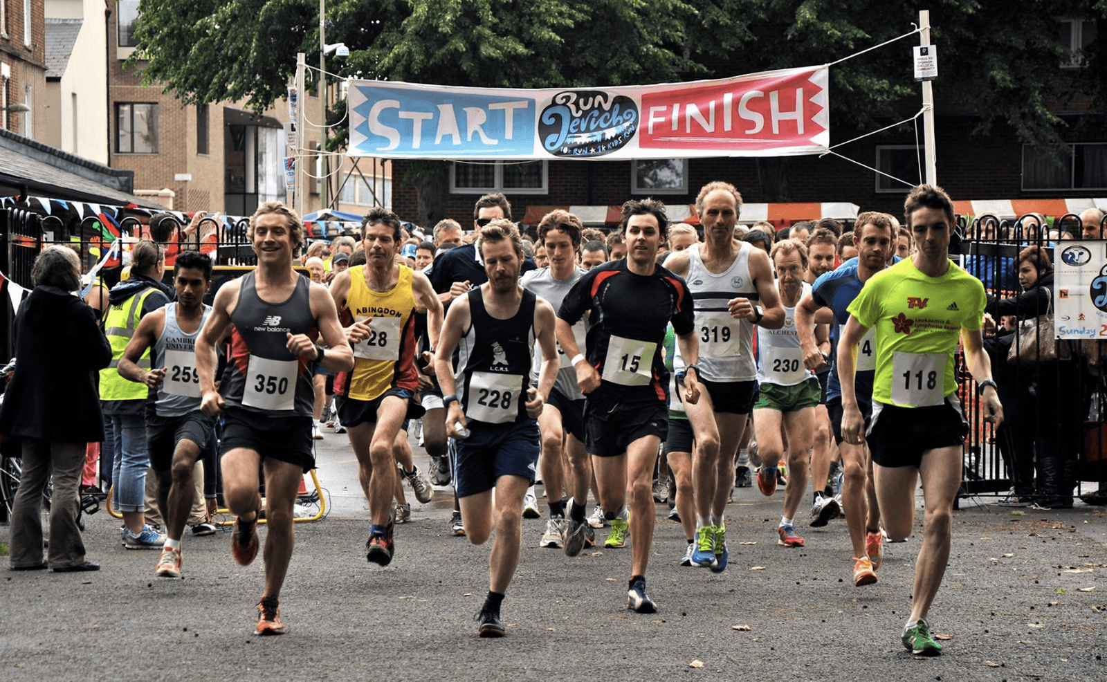 Runners starting a race beneath a banner that reads "START" and "FINISH" in a vibrant outdoor setting. They wear numbered bibs and athletic gear, surrounded by onlookers and trees in the background.