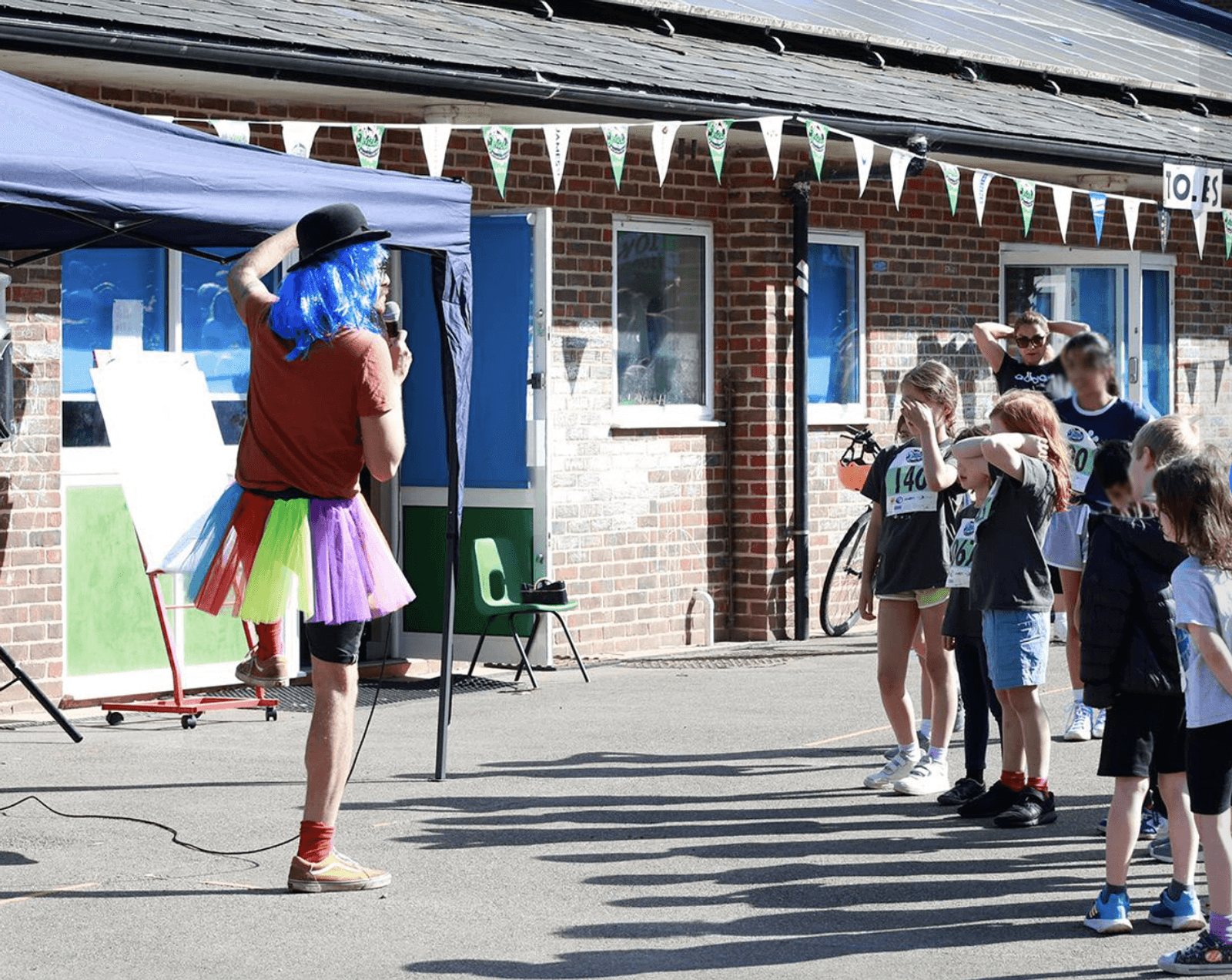 A person dressed in colorful clothing and a blue wig entertains a group of children outdoors. The scene is festive with bunting overhead. Some children have numbers on their shirts, possibly participating in an event. The building in the background is brick.