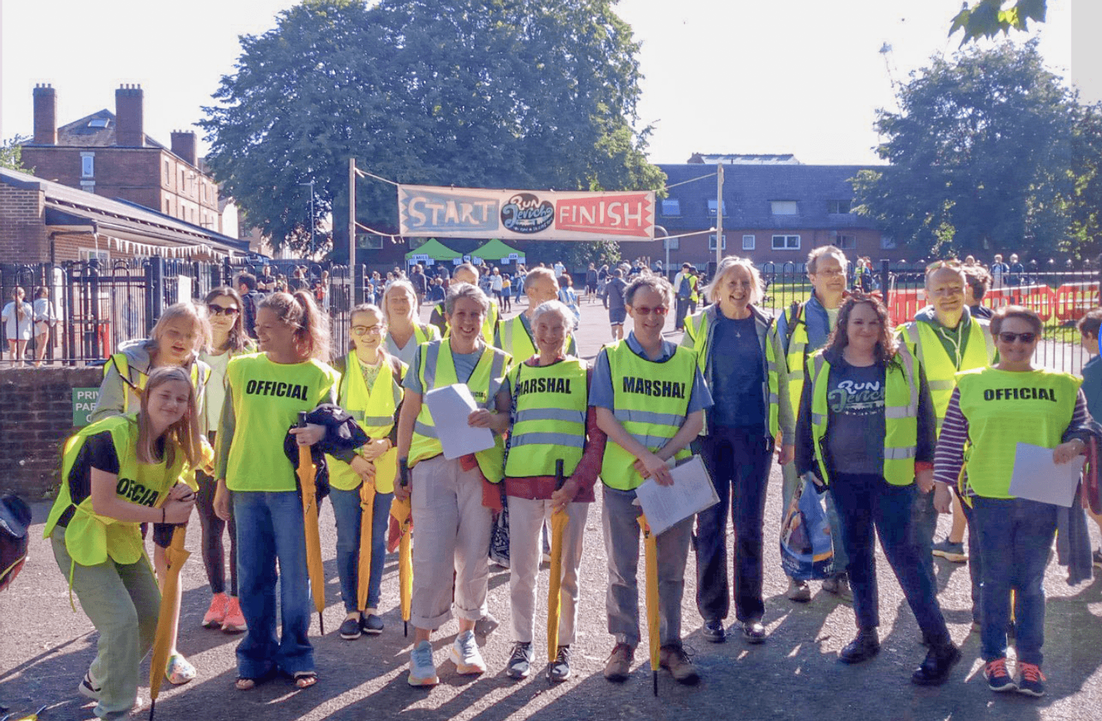A group of people, wearing bright yellow "Official" and "Marshal" vests, stand in front of a start/finish line banner at an outdoor event. They pose for the photo on a pathway, with trees and buildings in the background.