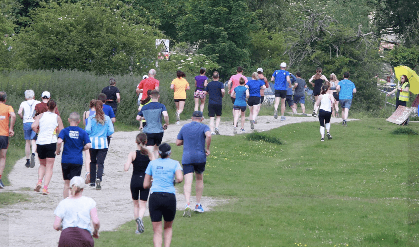 A group of people are running on a gravel path through a park. They are wearing athletic clothing in various colors. The path is surrounded by green grass and trees. The scene conveys a sense of community and outdoor activity.