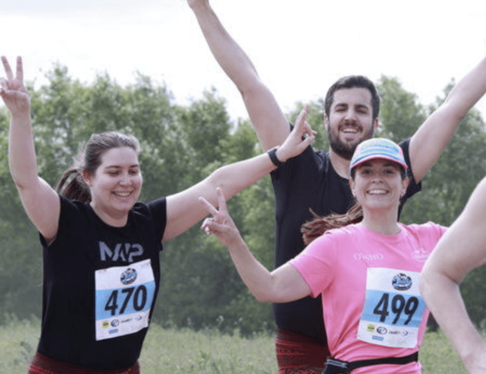 Three people joyfully pose with arms raised and smiling during a race. They wear numbered race bibs, with the woman on the left wearing black, the woman on the right in pink, and the man in the middle with a beard. Background shows greenery.
