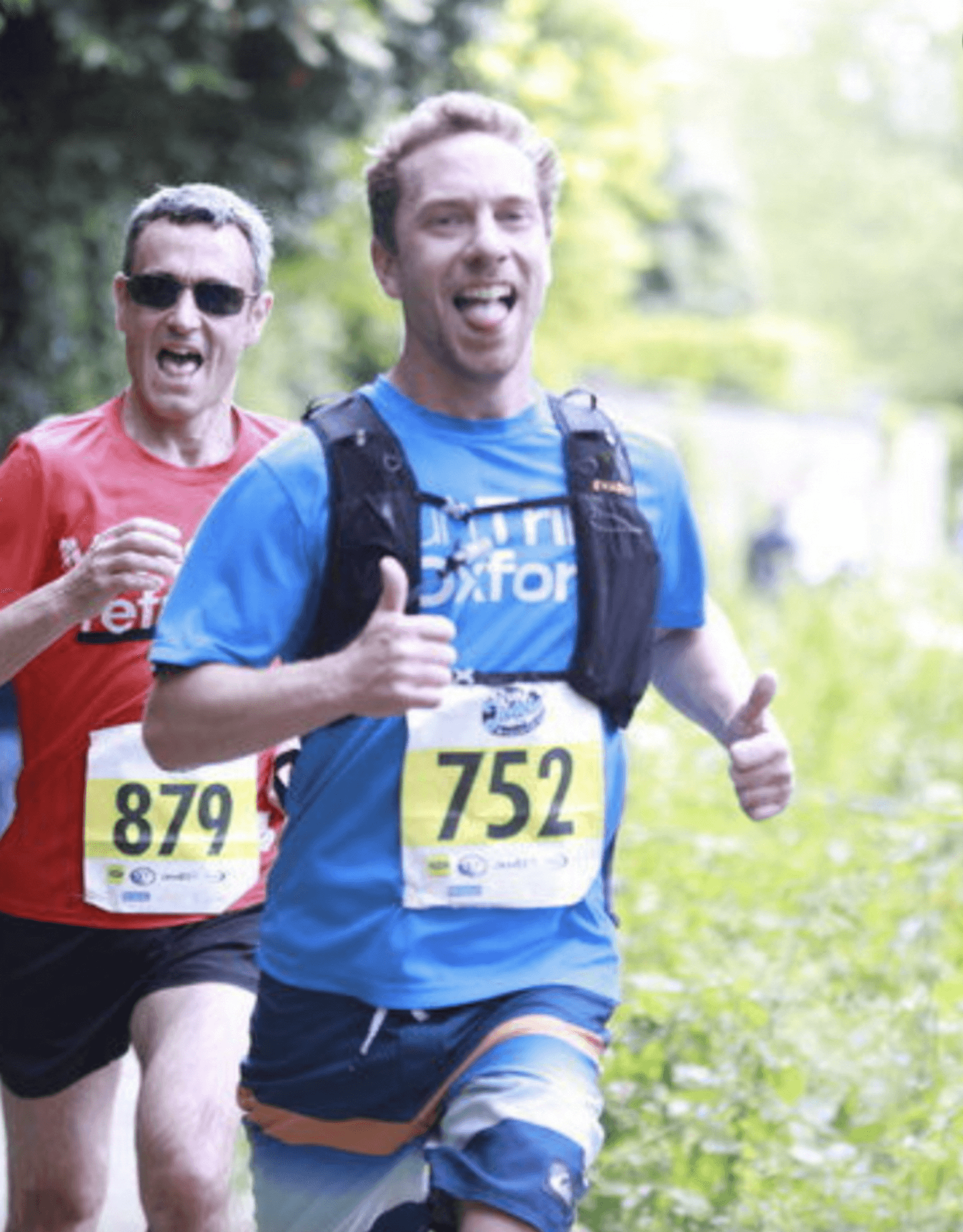 Two men are running a race in a lush, green outdoor setting. The man in the front is wearing a blue shirt and a race bib numbered 752, giving a thumbs-up and smiling. The man behind him is wearing a red shirt and sunglasses, bib number 879.