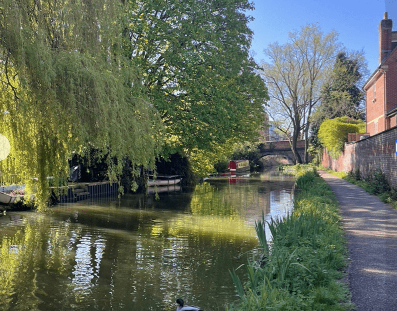 A serene canal scene with calm water reflecting a clear blue sky. Lush greenery lines the banks, including a weeping willow. A narrowboat is moored on the left, near an arched stone bridge. A red brick house stands on the right.