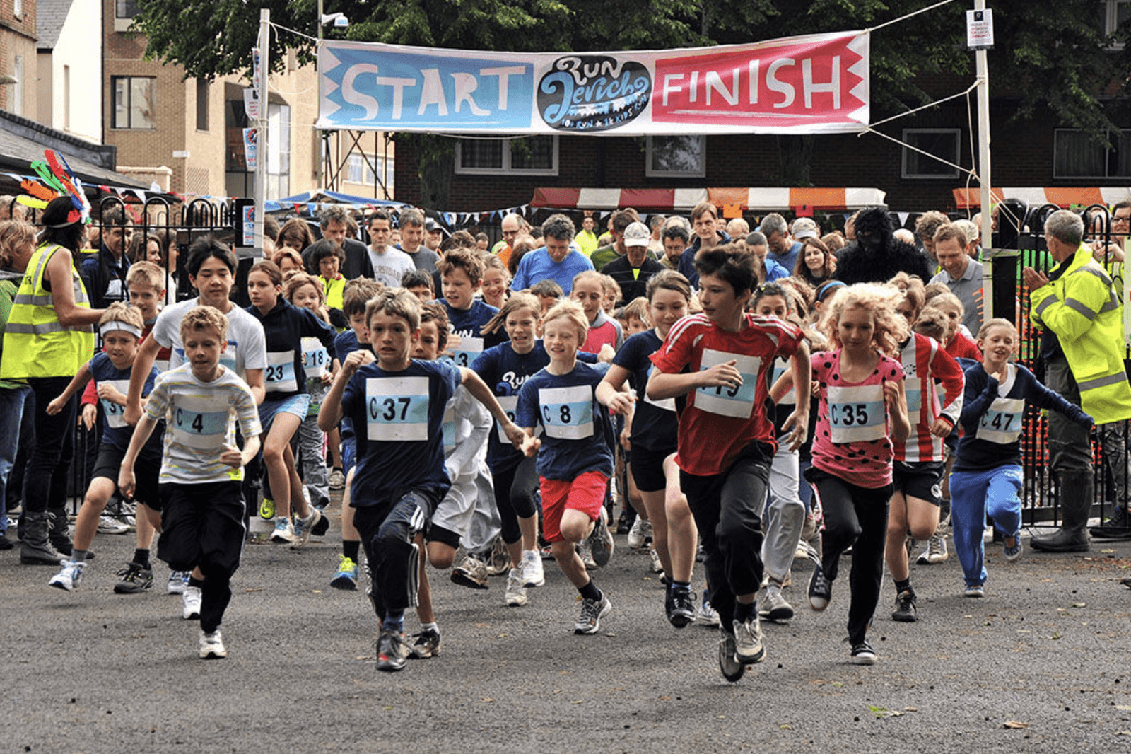 Children are energetically starting a race at an outdoor event. They're wearing numbered race bibs and running under a banner that reads "START" and "FINISH." A crowd of spectators and event staff are visible in the background.