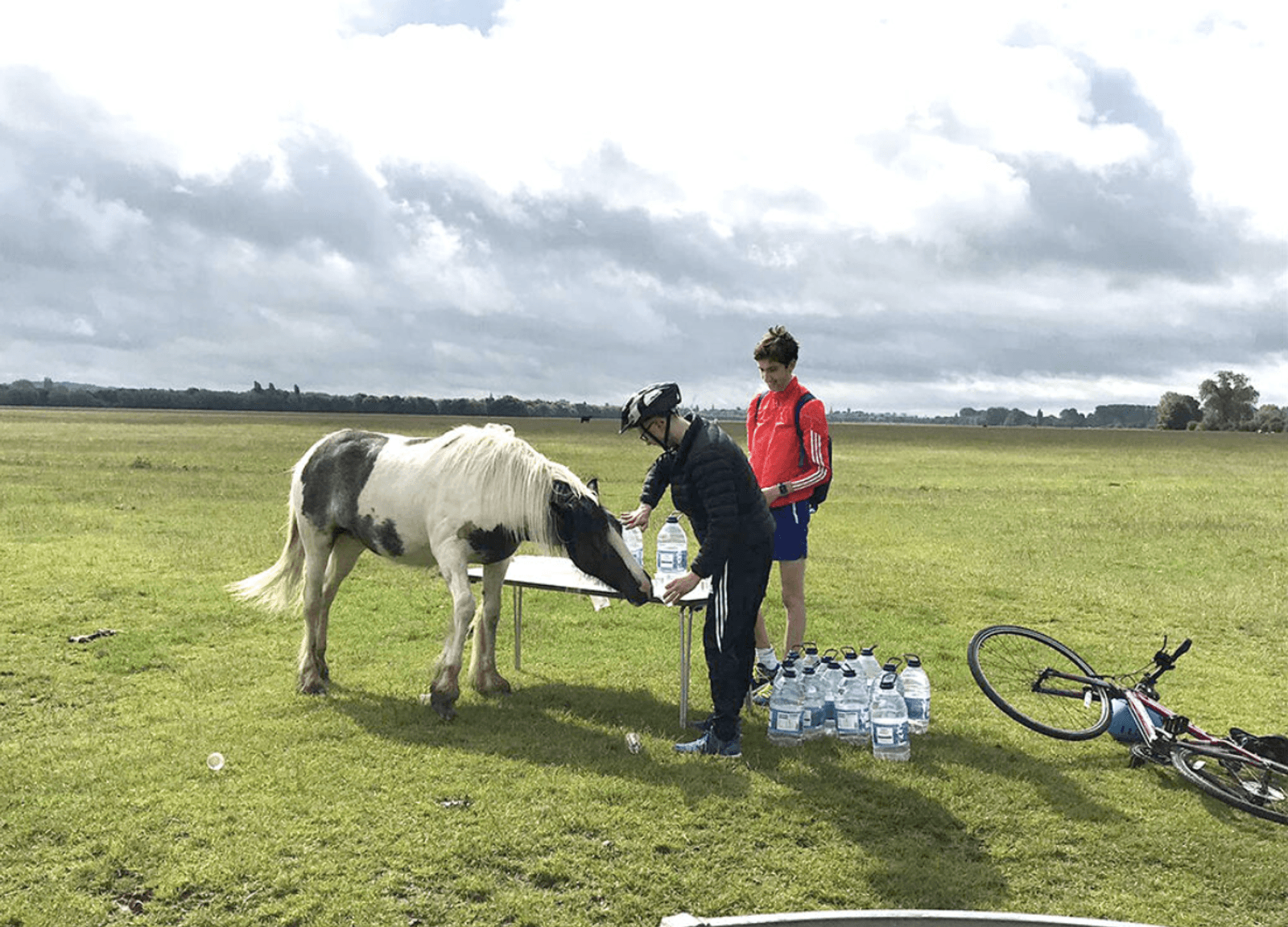 Cyclists stop at a table set up in a grassy field. One cyclist in a black helmet feeds a small white-and-black pony. Another cyclist in a red jacket stands nearby. Several bottles of water are on the table and ground, a bicycle lies nearby.