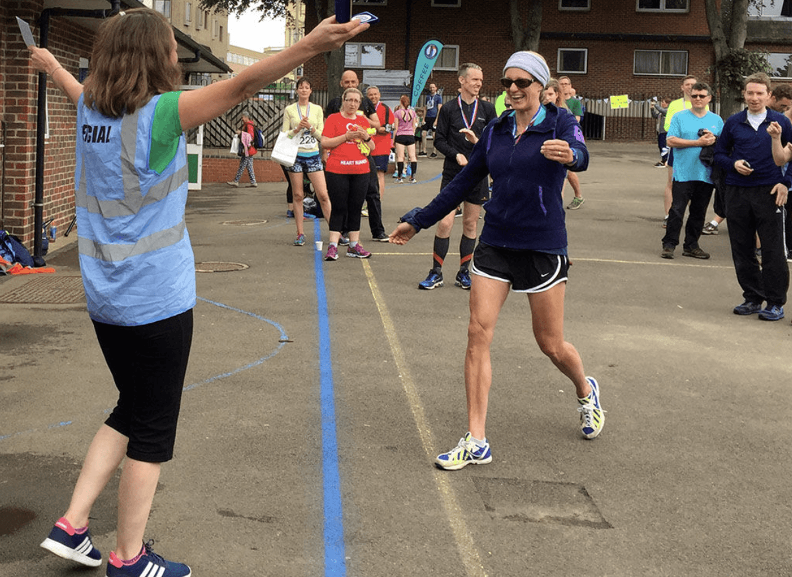 A woman wearing athletic gear approaches a finish line, arms outstretched, smiling, in a race event. A person in a "Marshall" vest welcomes her. Onlookers cheer in the background on a sunny day.