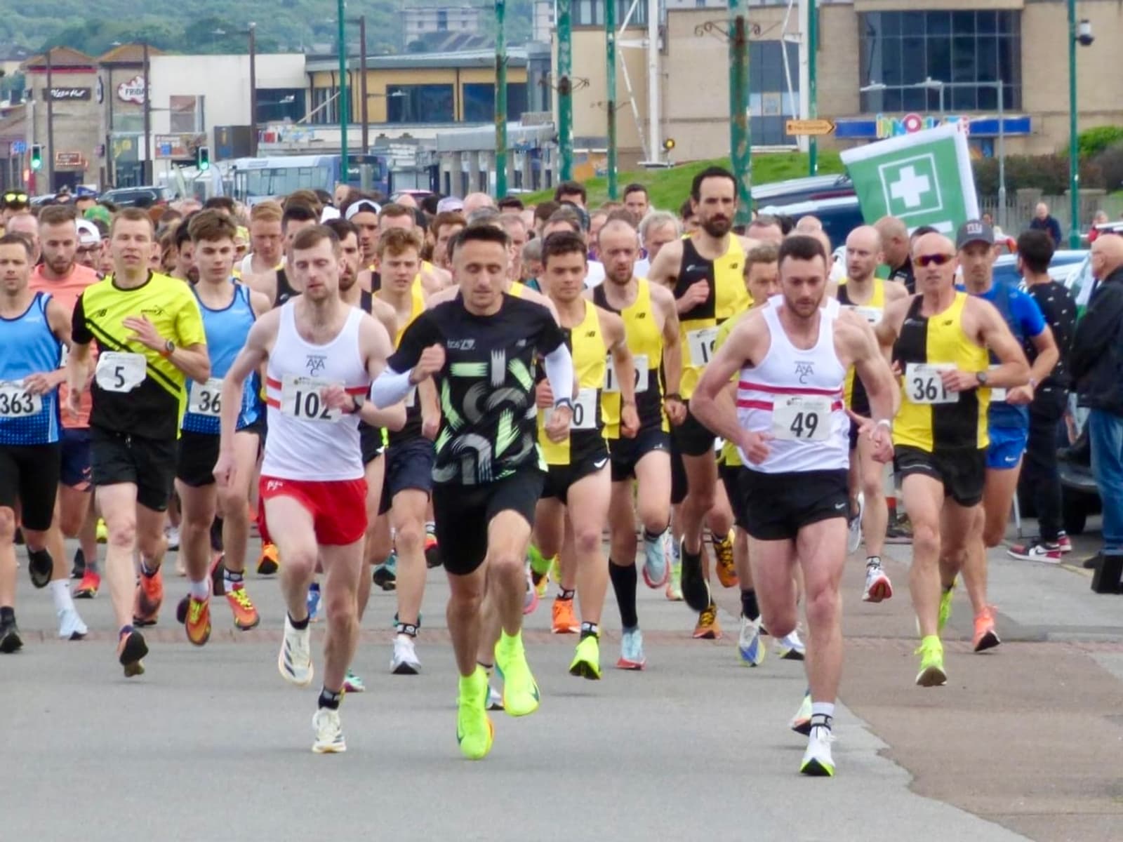 A large group of runners in colorful athletic wear is participating in a road race. They appear focused and energetic as they run along a street with various spectators watching. Numbered race bibs are visible on their shirts.