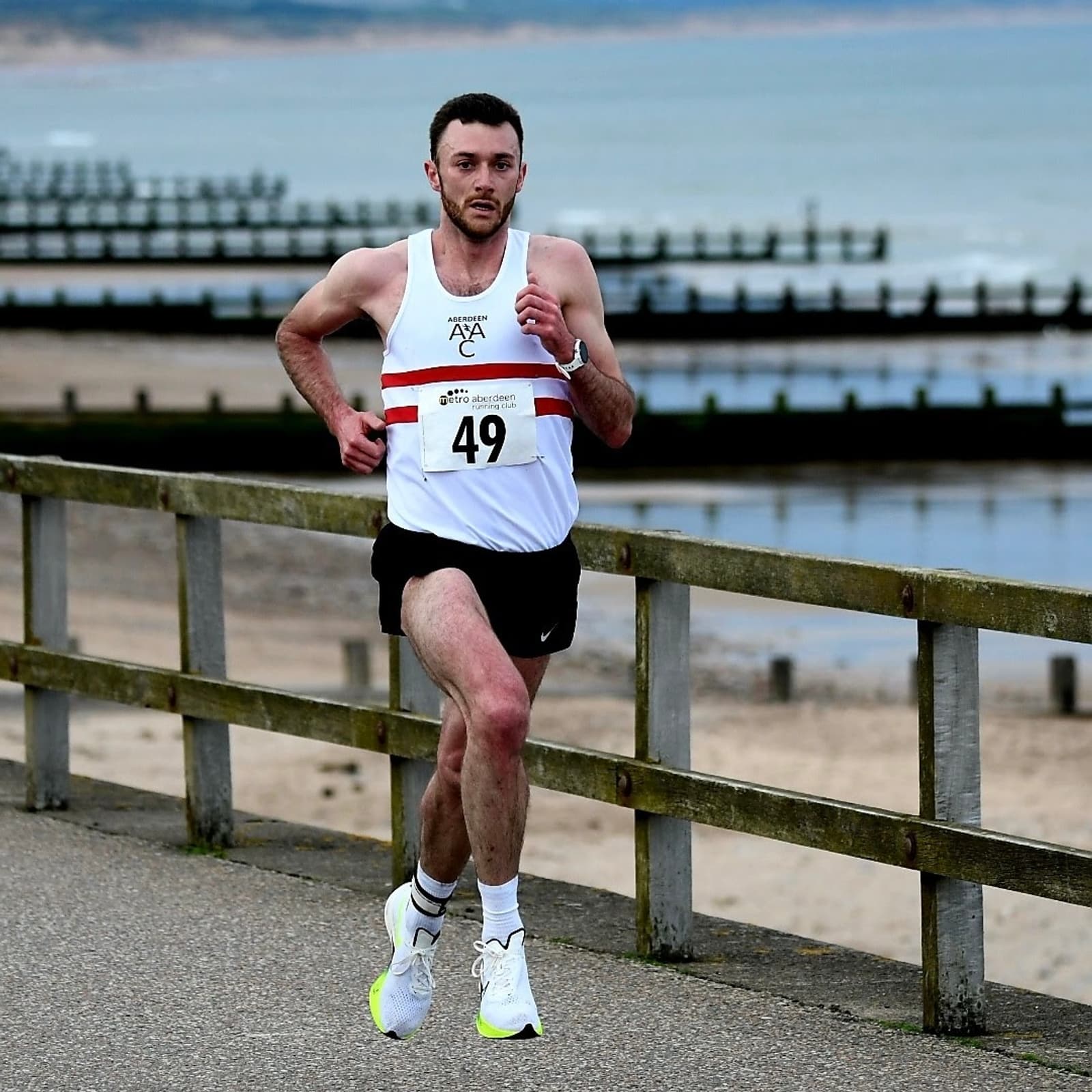 A runner in a white and red sleeveless shirt and black shorts is sprinting on a coastal path. He wears white shoes and has a number 49 bib. Wooden railings and a sandy beach with ocean waves are in the background.