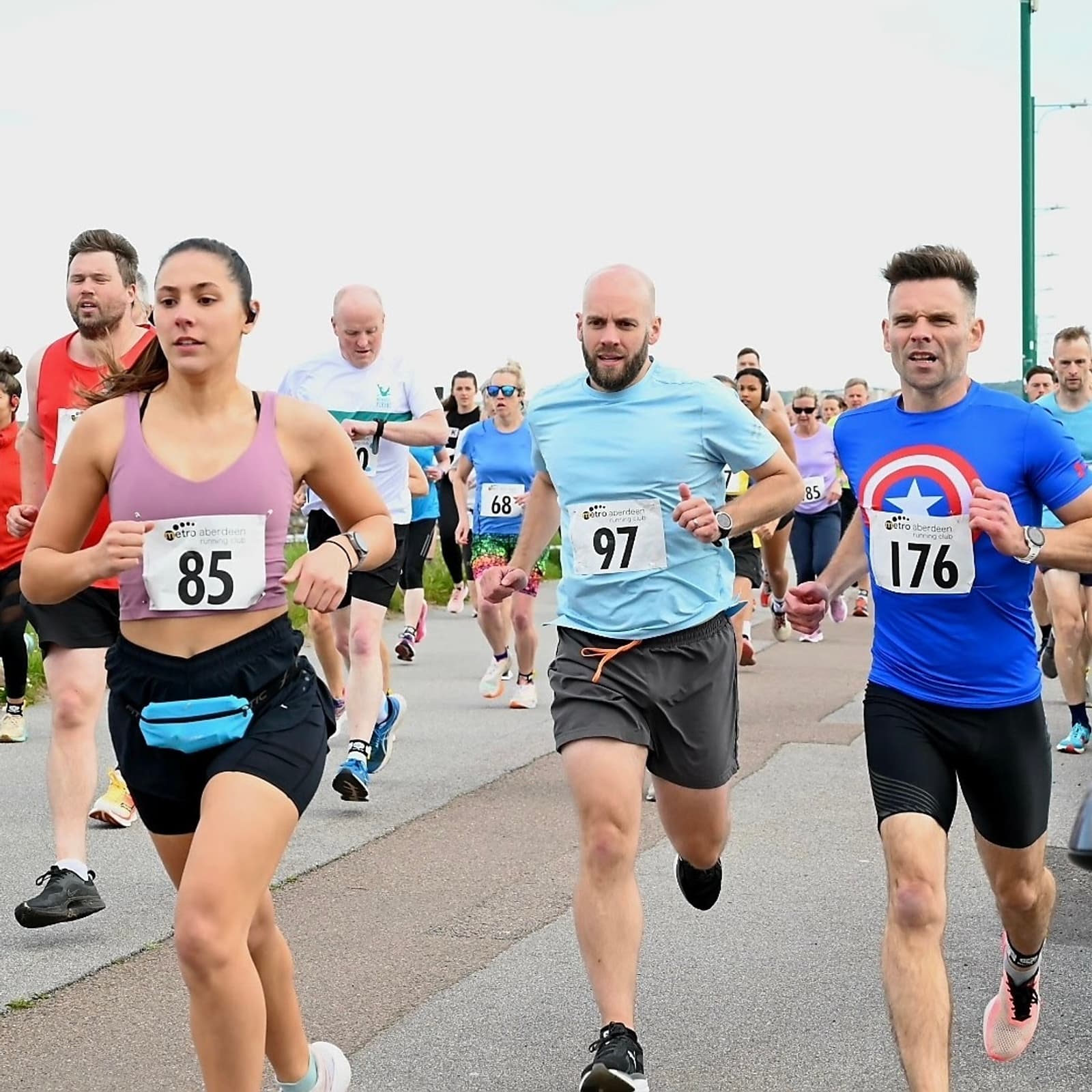 A group of people running a race on a paved road, wearing numbered bibs. The foreground shows a woman with bib 85 and two men with bibs 97 and 176. They are focused and determined, with other runners visible in the background.