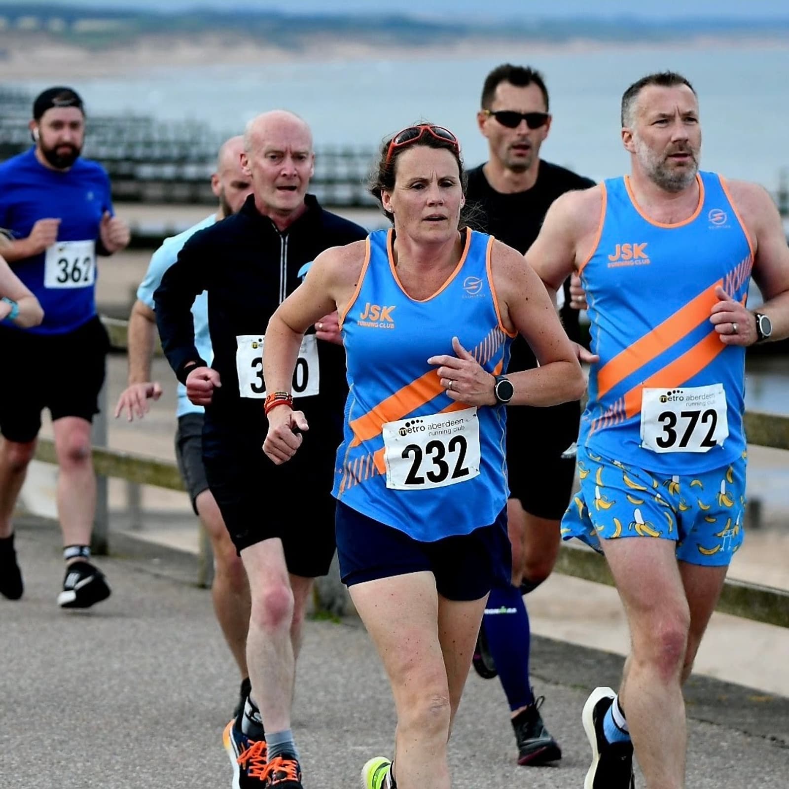 Runners participate in a road race along a seaside path. A woman in the foreground, wearing a blue and orange outfit, is focused on the race. Other runners, including men in similar attire, are close behind. The ocean is visible in the background.