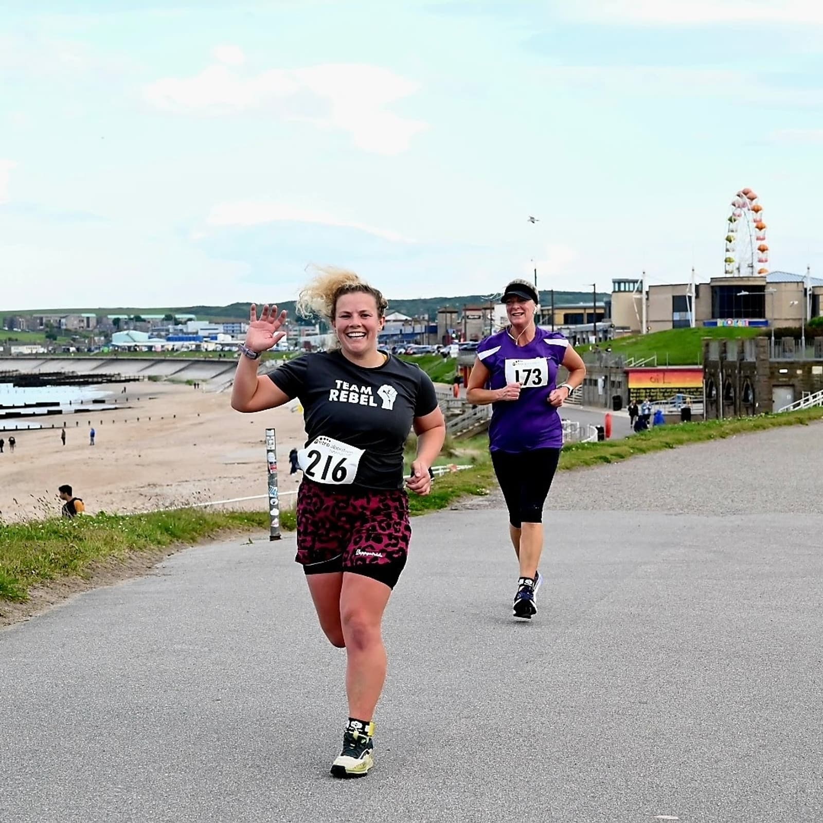 Two runners on a seaside path during a race. The runner in front wears a gray shirt and leopard print shorts, smiling and waving. The second runner wears a purple shirt. A ferris wheel and beach are visible in the background.