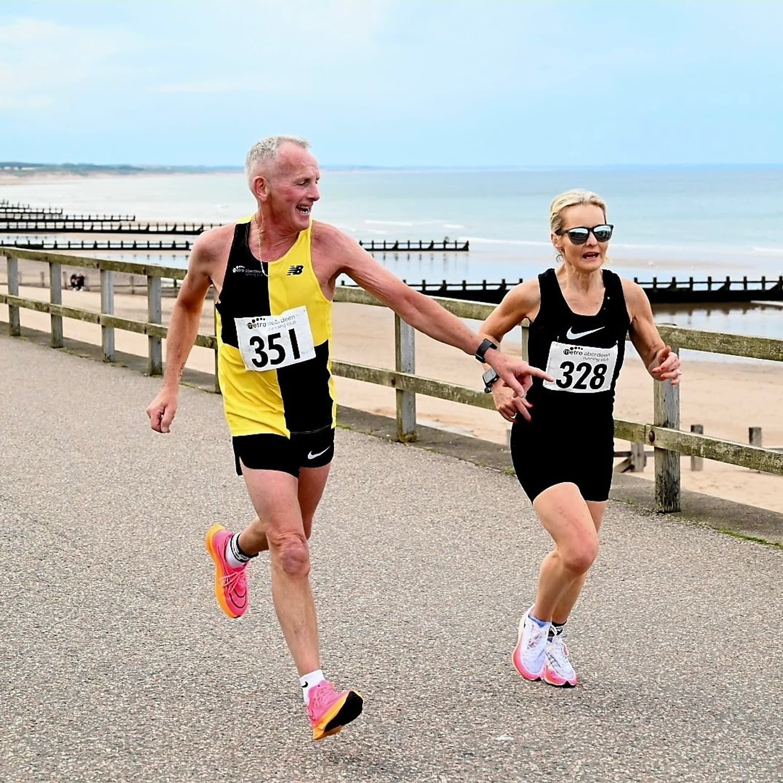 Two runners holding hands during a race by the beach. The man, in a yellow and black outfit with bib number 351, smiles as he runs alongside a woman in black with bib number 328. The ocean and a clear sky are visible in the background.