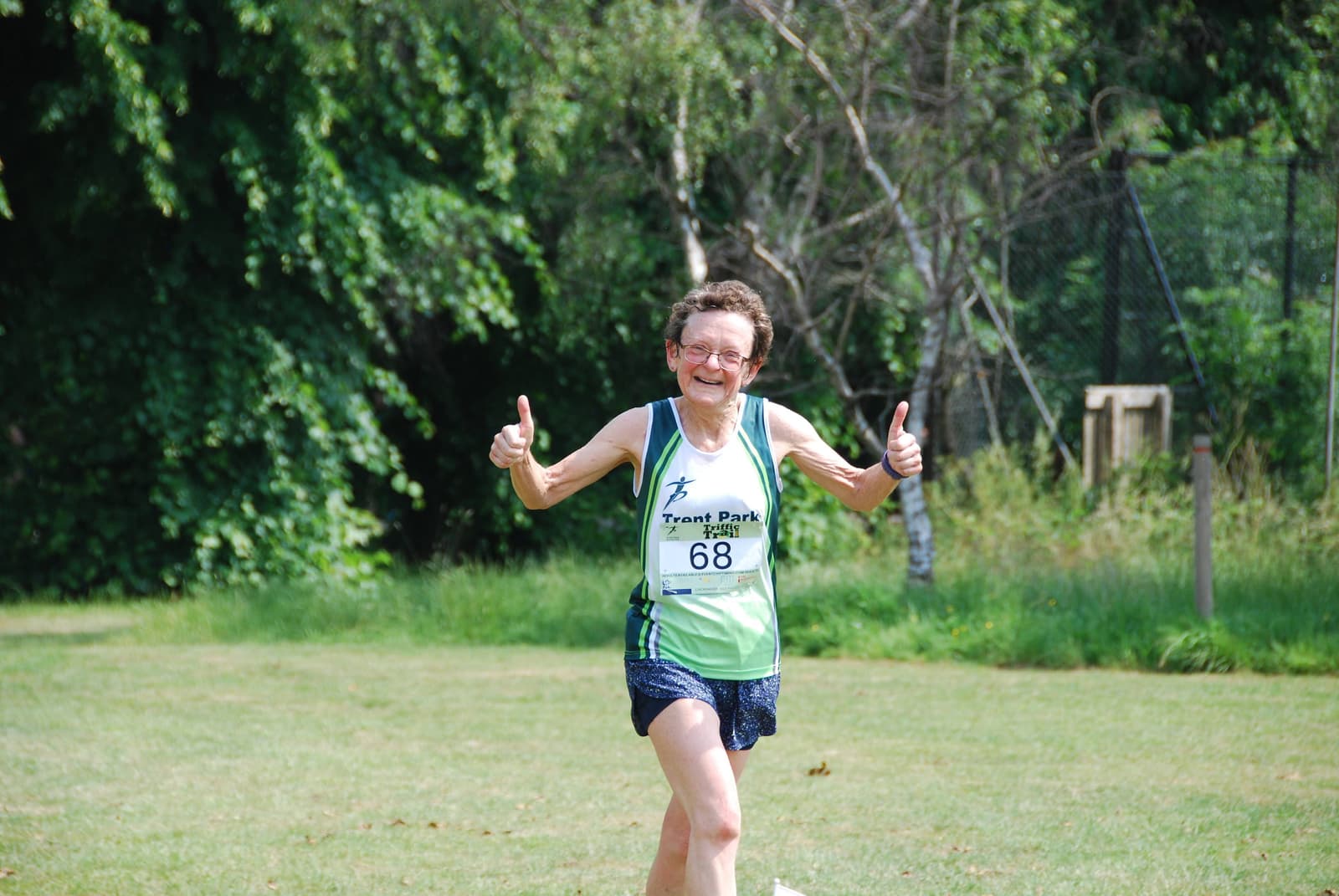 Woman wearing a green tank top with a number "68" and shorts, giving two thumbs up while running on a grassy path. Trees and a wire fence are in the background, suggesting a park setting.