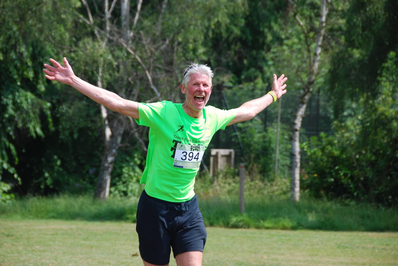 An older man is running outdoors on grass with arms outstretched, wearing a bright green shirt and black shorts. He is smiling, wearing a race number "394," and is surrounded by greenery and trees in the background on a sunny day.