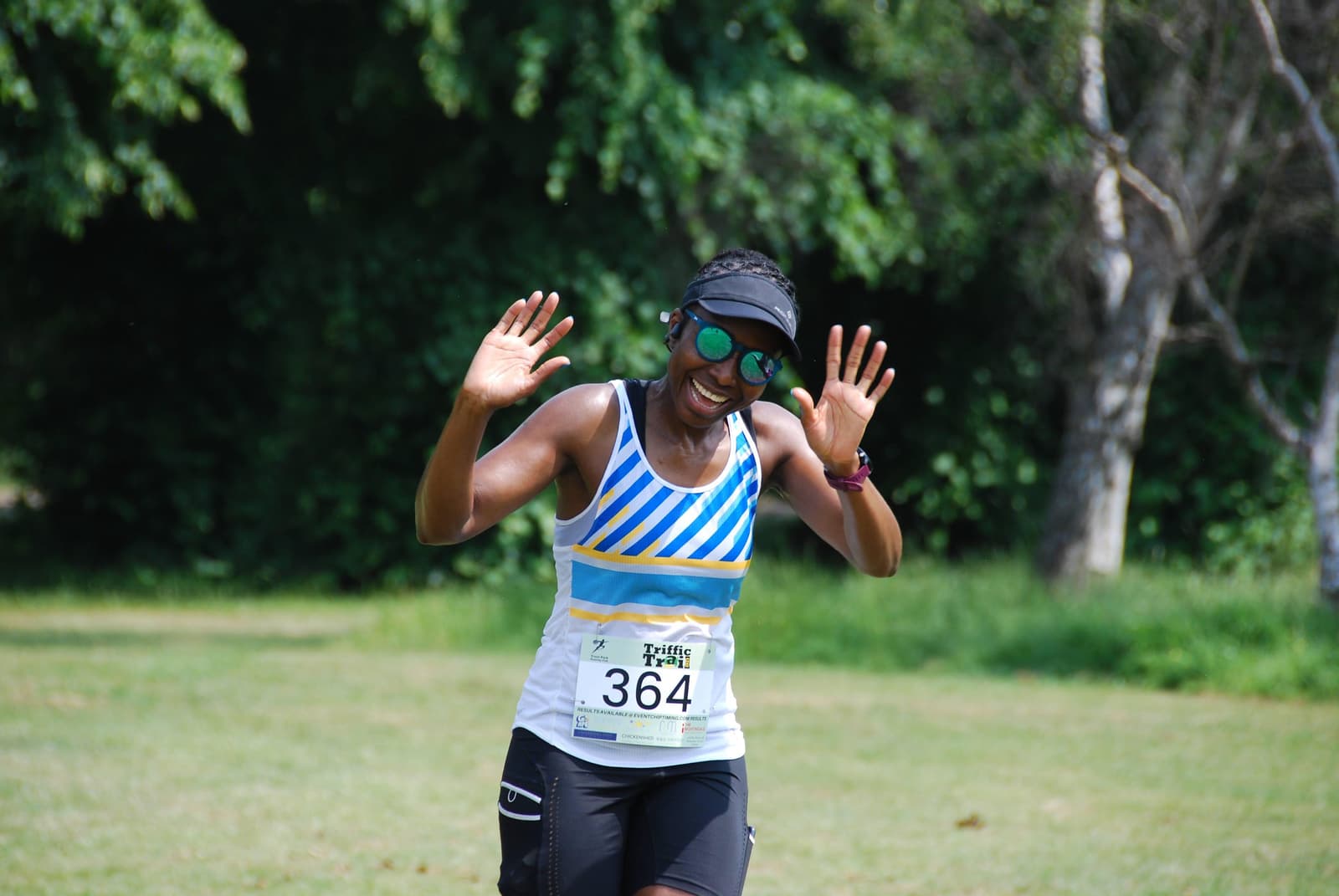 A runner with sunglasses and a colorful tank top smiles and raises their hands in victory while jogging on a grassy field, with trees in the background. They are wearing a race number on their waist.