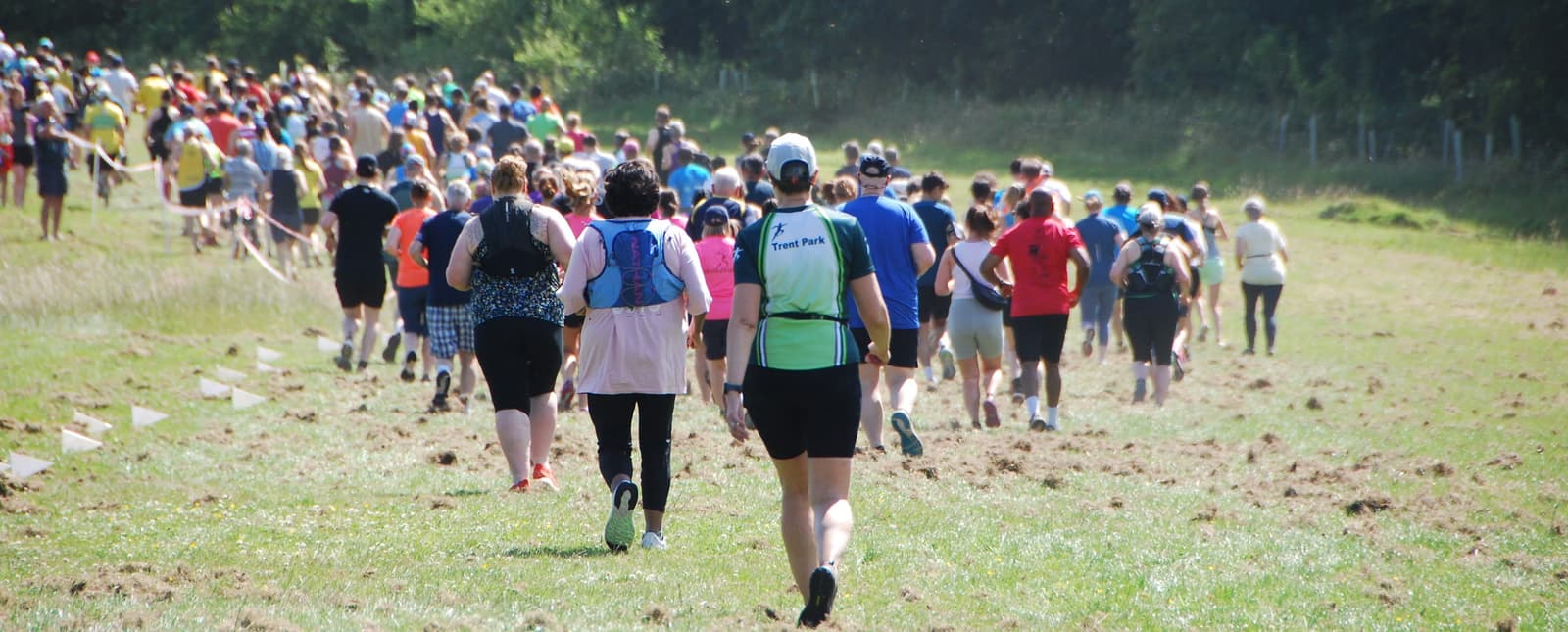 A large group of people participating in an outdoor race or fun run on a grassy field. They are wearing casual athletic gear, including shorts, t-shirts, and caps. The background features trees and a bright, sunny sky.