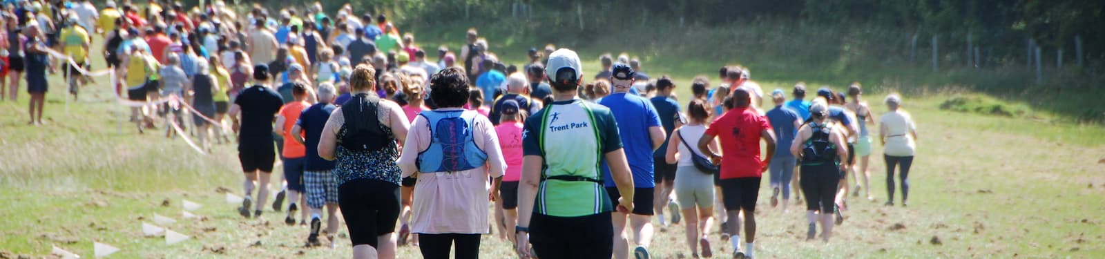 A large group of people in athletic clothing is walking and running together across a grassy outdoor trail, surrounded by trees and fenced areas, during what appears to be a race or organized event.