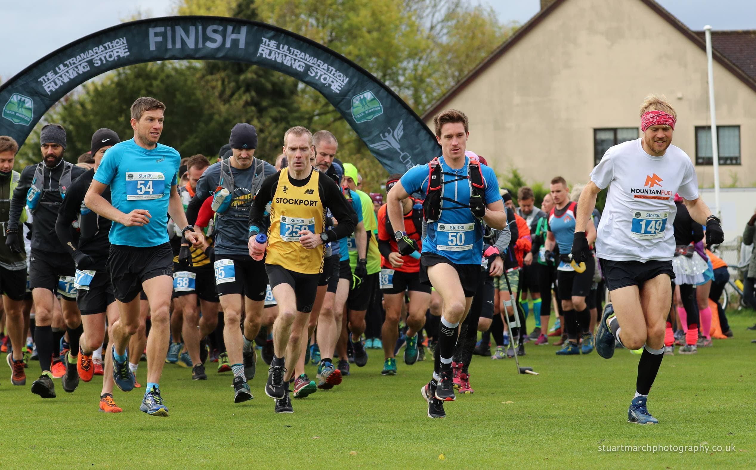 A group of runners in athletic gear start a race on grass, passing under a black inflatable arch that says "FINISH." Spectators and a house are visible in the background.