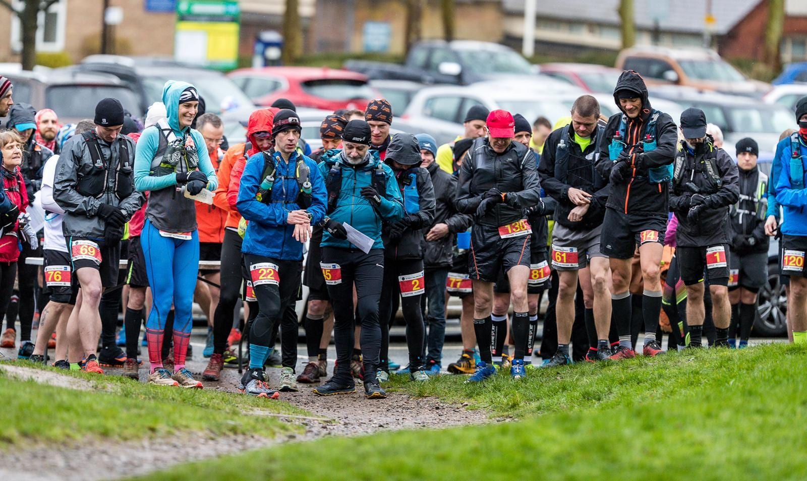 A group of runners in athletic gear gather at a muddy start line during a race, many wearing jackets, backpacks, and hats, with numbered bibs visible, and cars parked in the background.
