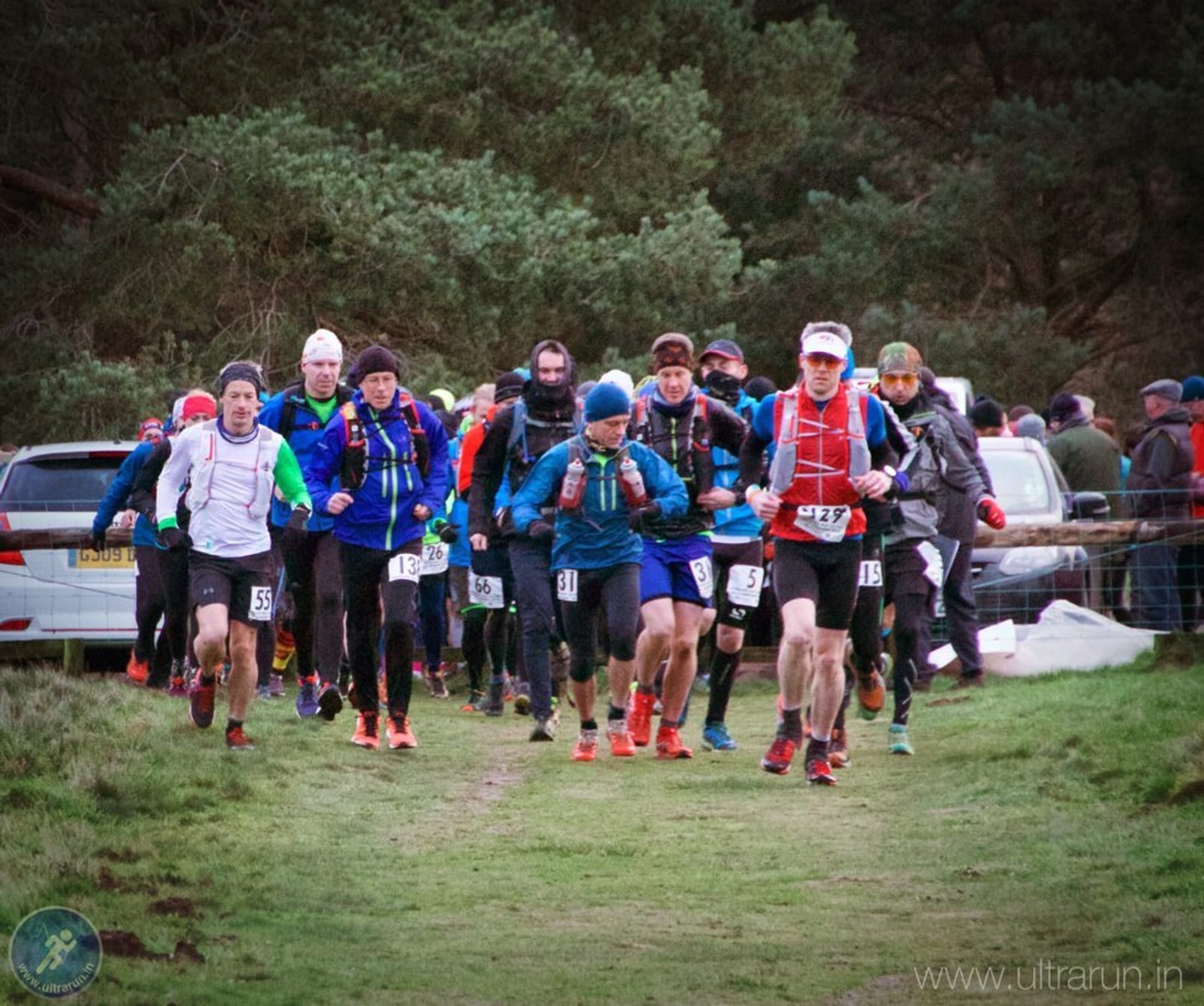 A group of runners dressed in outdoor gear start a trail race on a grassy path, surrounded by trees and spectators near parked cars. The atmosphere is energetic and focused.