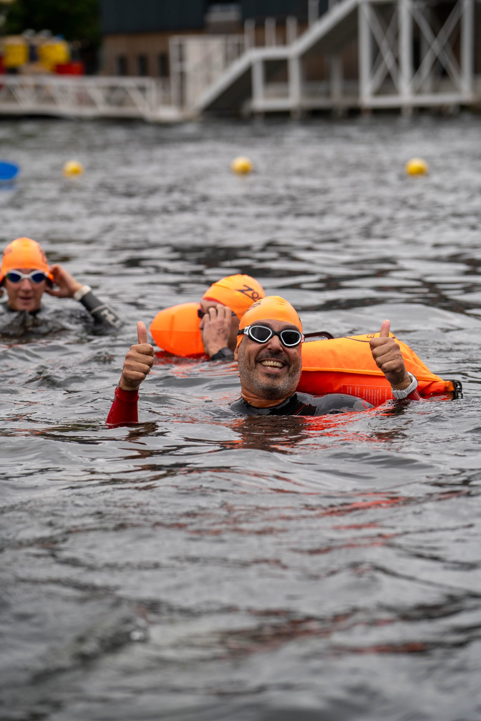 A group of swimmers wearing orange swim caps and goggles float in open water, with one smiling man in the foreground giving two thumbs up. Orange safety buoys are visible behind them.