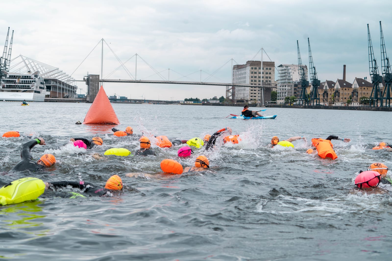 A large group of swimmers wearing colorful swim caps and safety buoys participate in an open water race in a harbor, with a kayaker nearby and industrial buildings, cranes, and a bridge in the background.