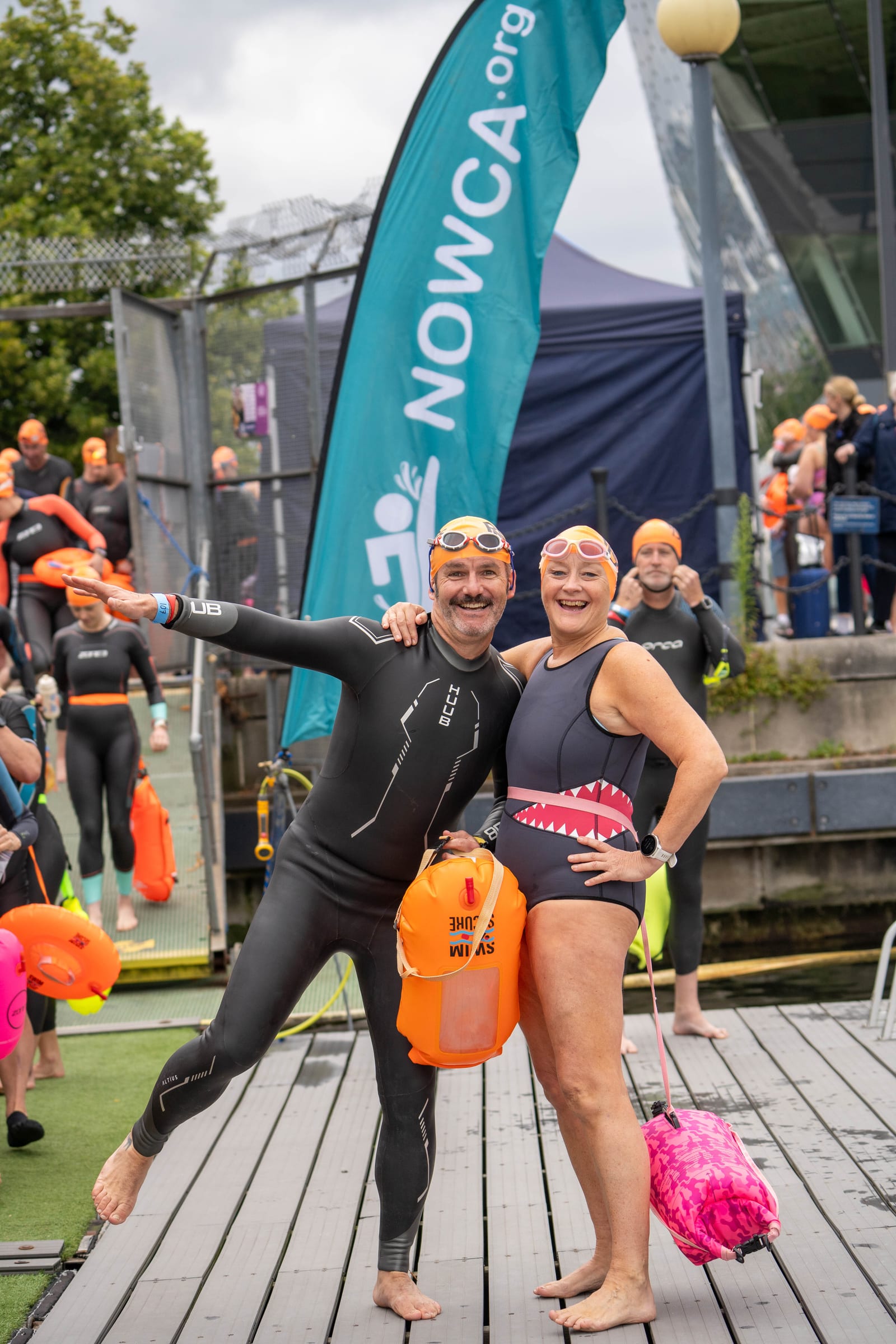 Two swimmers in wetsuits and orange swim caps smile and pose together on a dock, holding tow floats. Other swimmers and a NOWCA.org banner are visible in the background. It appears to be an outdoor swimming event.