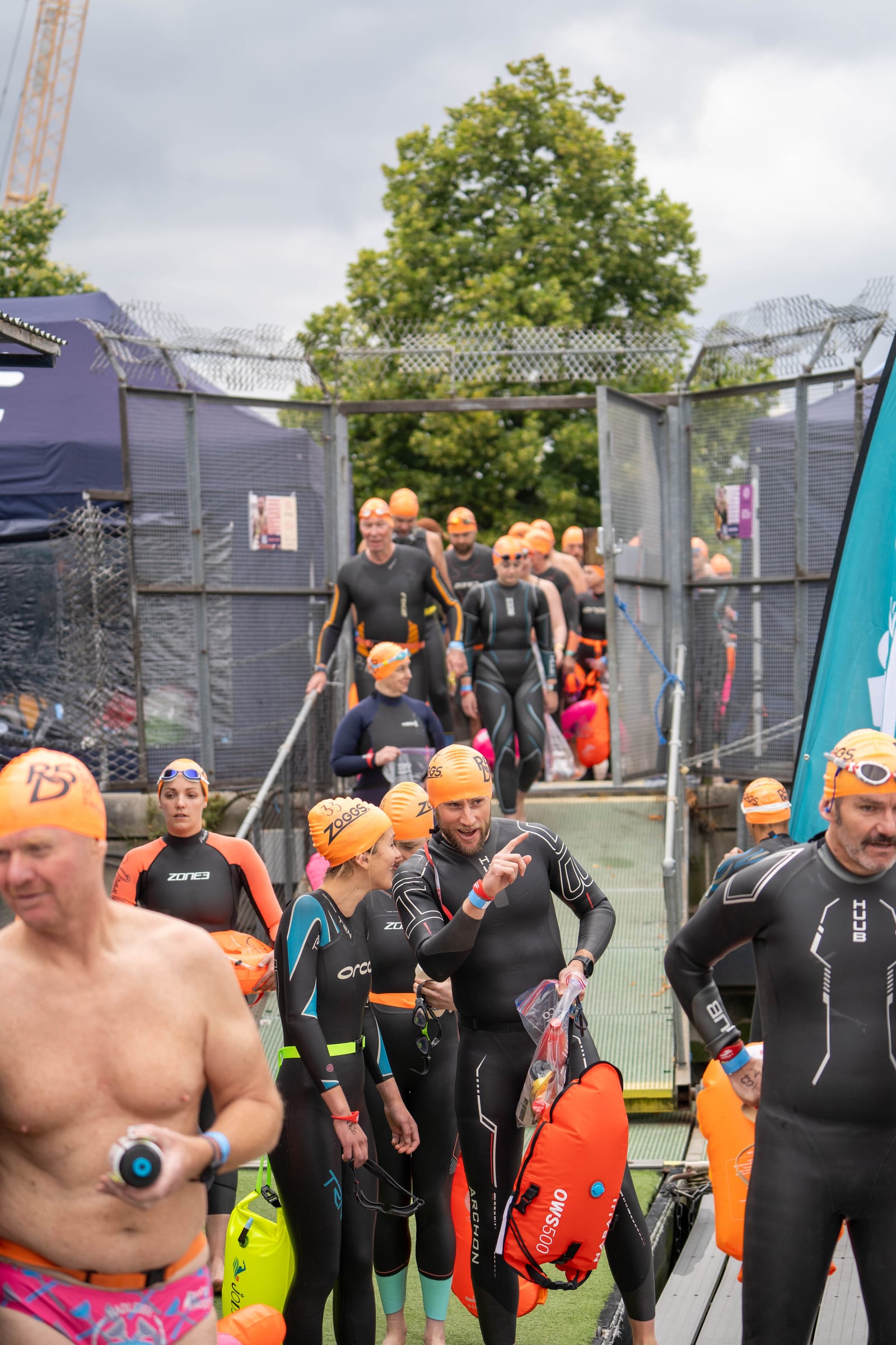 A group of people in wetsuits and orange swim caps walk along a ramp, holding orange floatation devices, preparing for an outdoor swim event. Trees and tents are visible in the background.