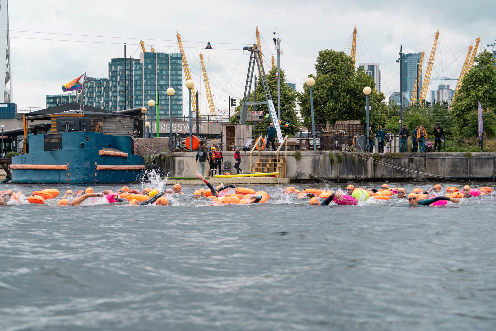 A group of swimmers with brightly colored floats participate in an open water event near a dock, with spectators watching and city buildings and cranes visible in the background.