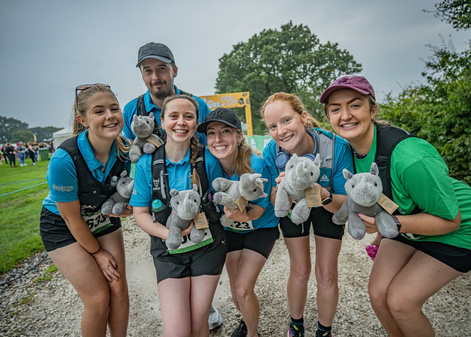 Six smiling people dressed in sporty outfits pose together outdoors, each holding a gray stuffed rhinoceros toy. There are trees and a yellow event archway in the background. The setting appears to be a fun run or charity event.