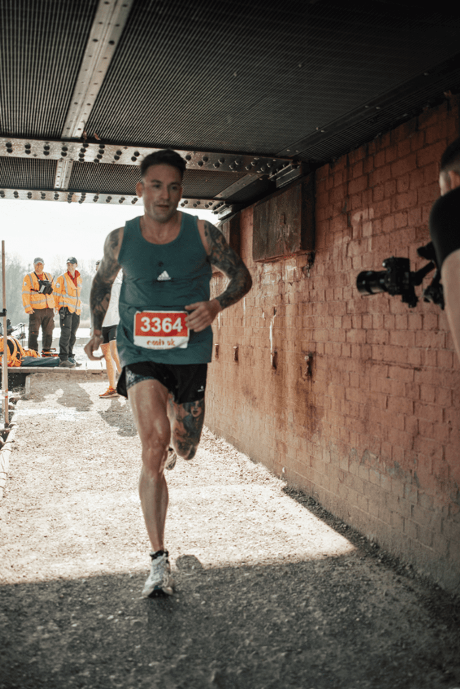 A male runner with tattoos, wearing a blue tank top and race bib 3364, runs through a tunnel in a race as officials and a cameraman look on in the background. Sunlight streams in from the tunnel entrance.
