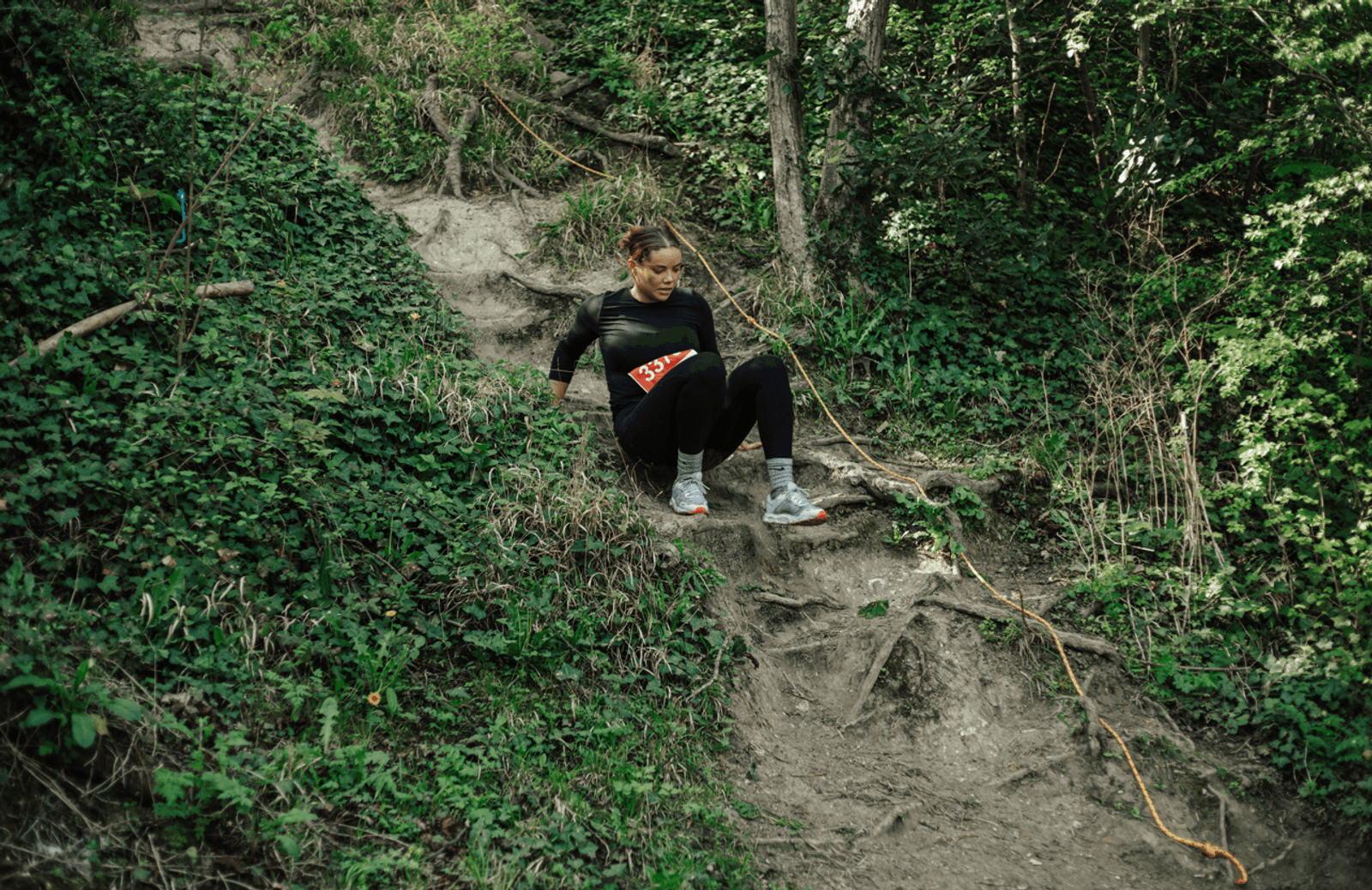 A person wearing athletic clothing and a race bib carefully climbs down a steep, muddy trail surrounded by green foliage in a forest.