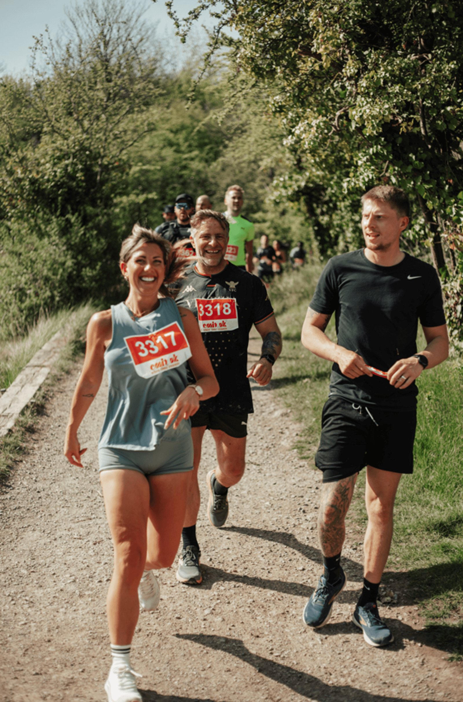 Three people with race numbers run and smile on a sunny dirt trail lined with trees and grass, surrounded by other runners in the background. The atmosphere is energetic and cheerful.