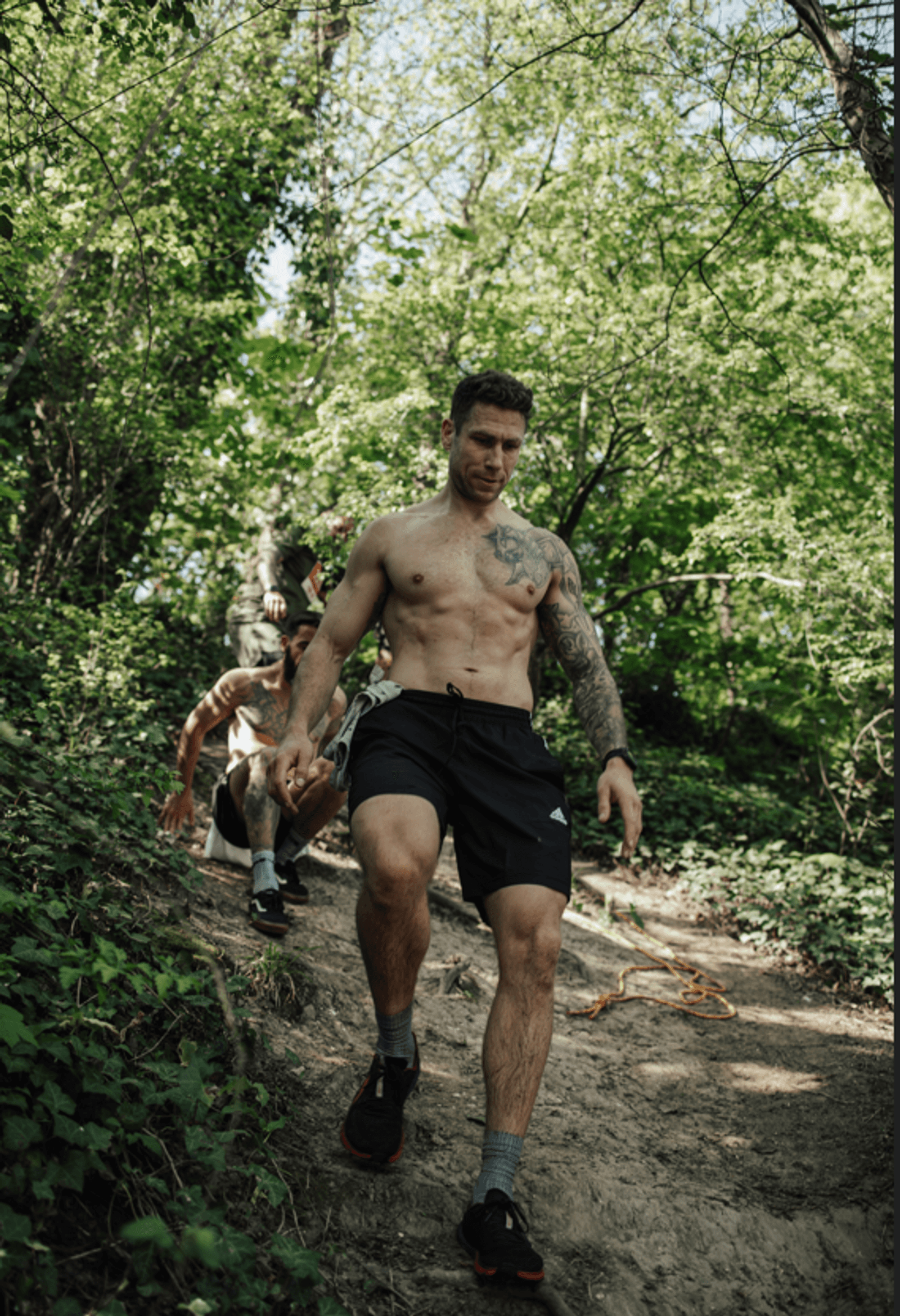 A shirtless man with tattoos and wearing black shorts hikes down a dirt path in a lush, green forest, followed by two other people in the background.