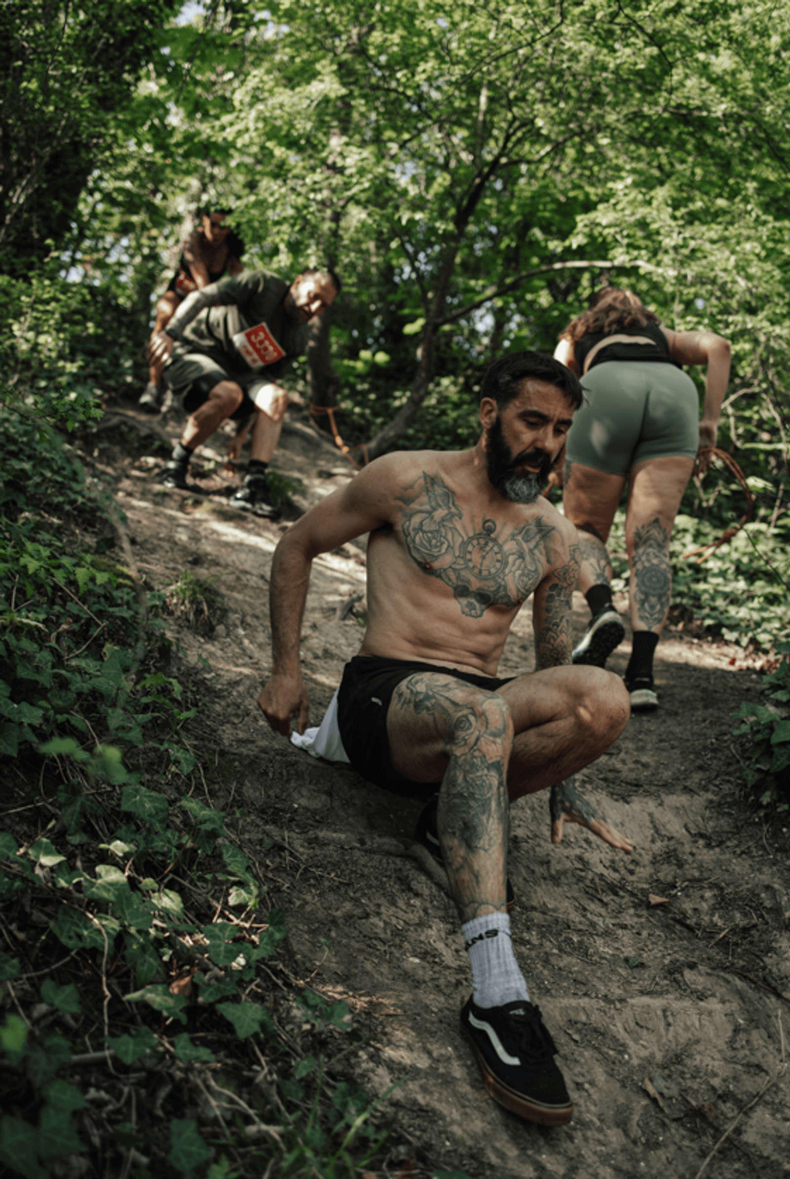 A shirtless, tattooed man slides down a steep, dirt path in a forest, followed by three people climbing or sliding down the same slope, surrounded by green foliage.