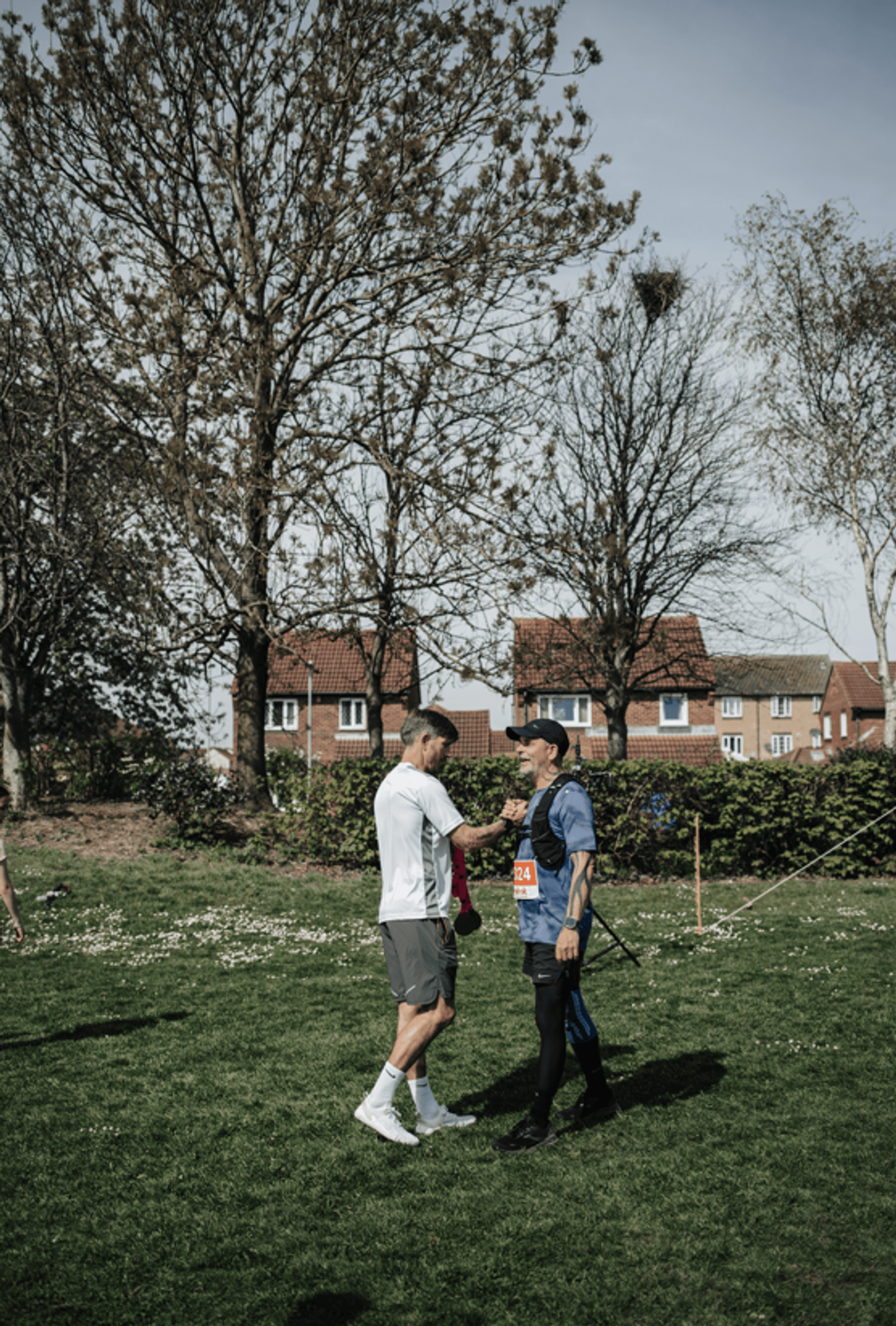 Two men stand facing each other on a grassy field, one in sportswear and running shoes, the other wearing a numbered race bib. They appear to be congratulating or greeting each other outdoors with houses in the background.